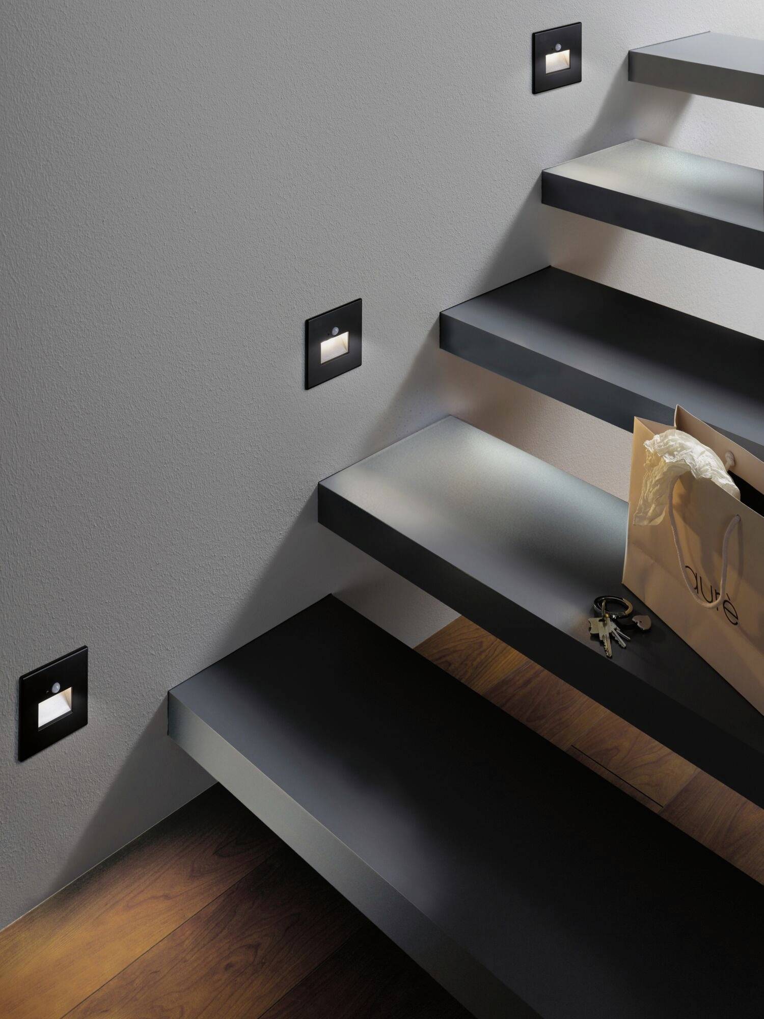 Escalier noir et gris avec des appliques murales modernes. Un sac en papier et des clés sont posés sur les marches. Design minimaliste.