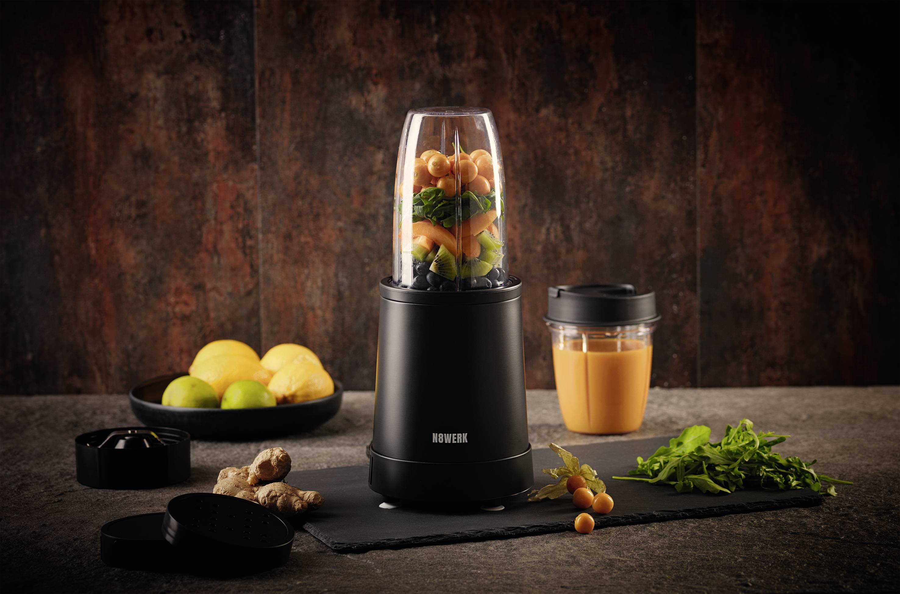 Mixeur compact rempli de légumes à côté d'un verre de jus fraîchement préparé, entouré de gingembre, de citrons et d'herbes sur un fond sombre.