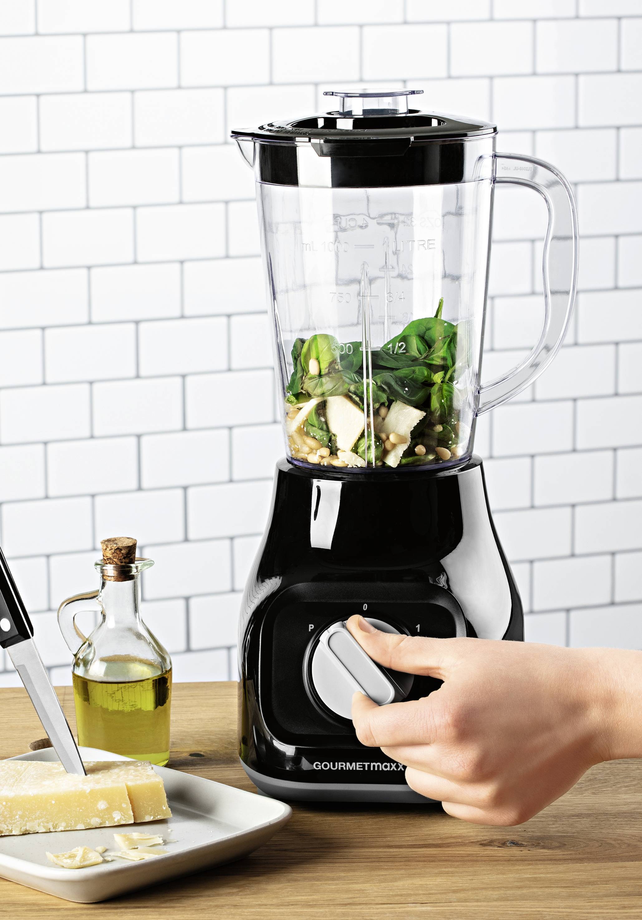Une main tourne le bouton d'un mixeur noir rempli d'épinards, de noix et de fromage. À côté se trouvent de l'huile et un bloc de fromage.
