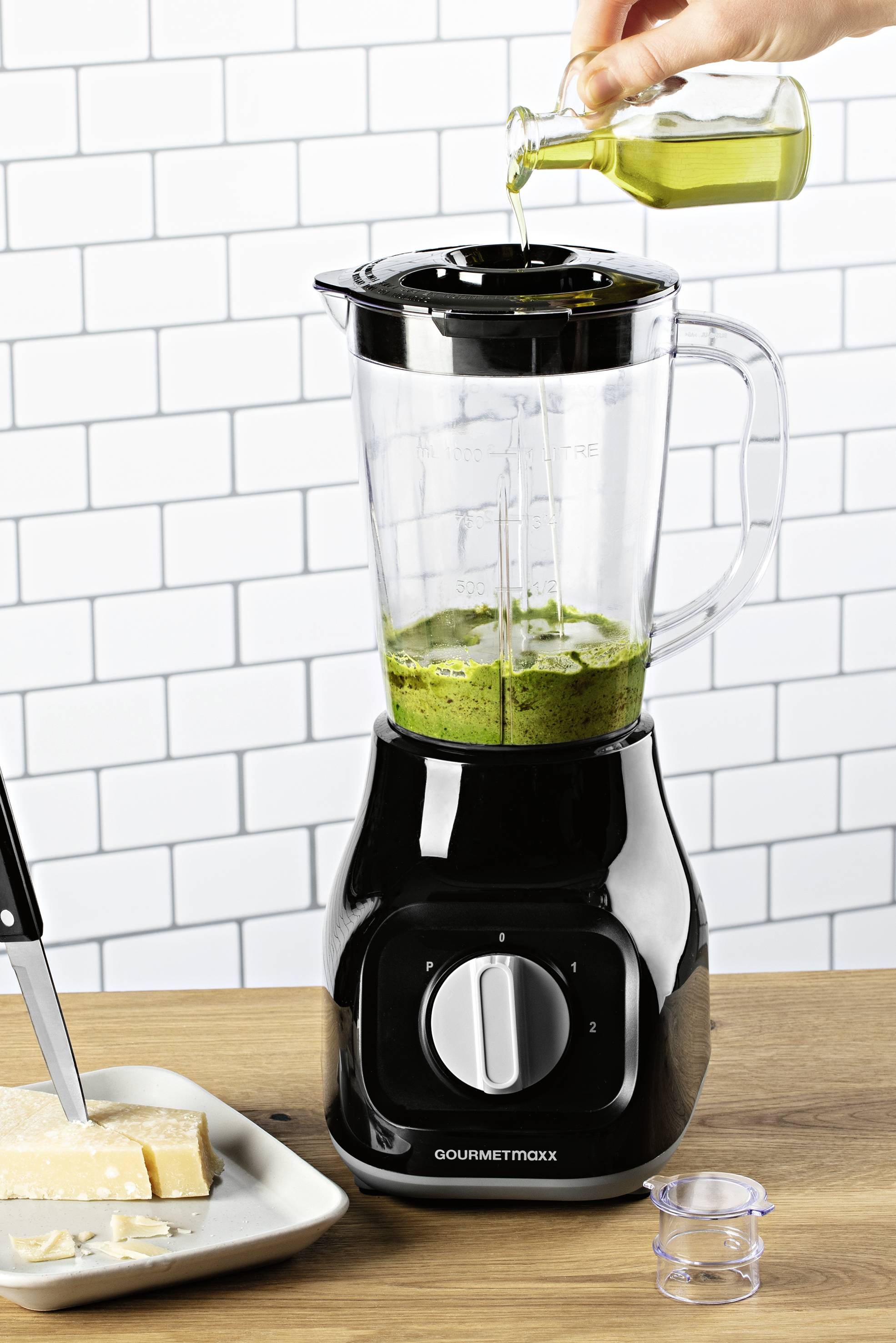 Une personne verse de l'huile dans un mixeur contenant un mélange vert. À côté, une assiette avec des cubes de fromage et un couteau sur une table en bois.