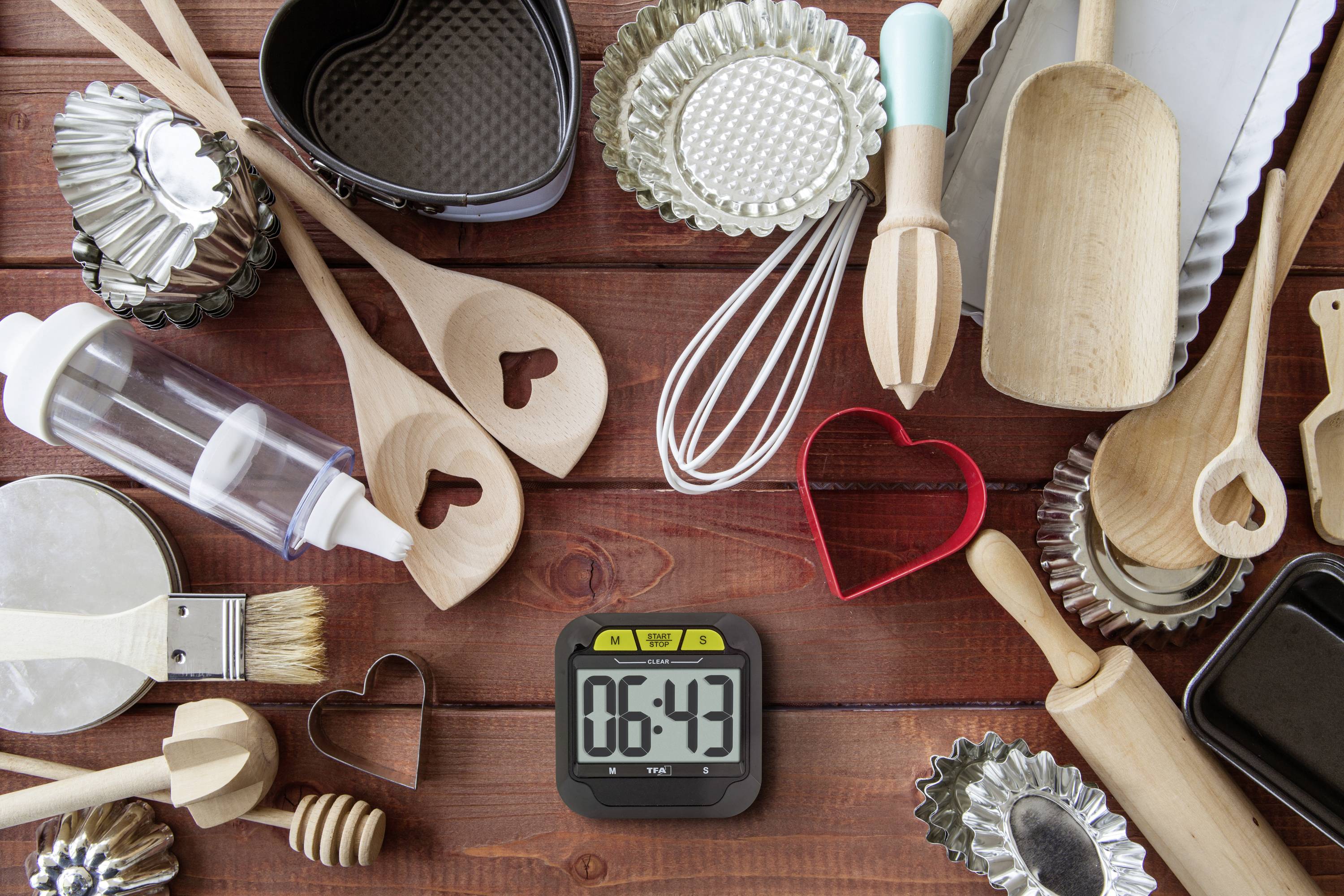 Divers ustensiles de pâtisserie comme des cuillères en bois, des emporte-pièces et une spatule à pâtisserie sont posés sur une table. Un minuteur affiche 06:43.