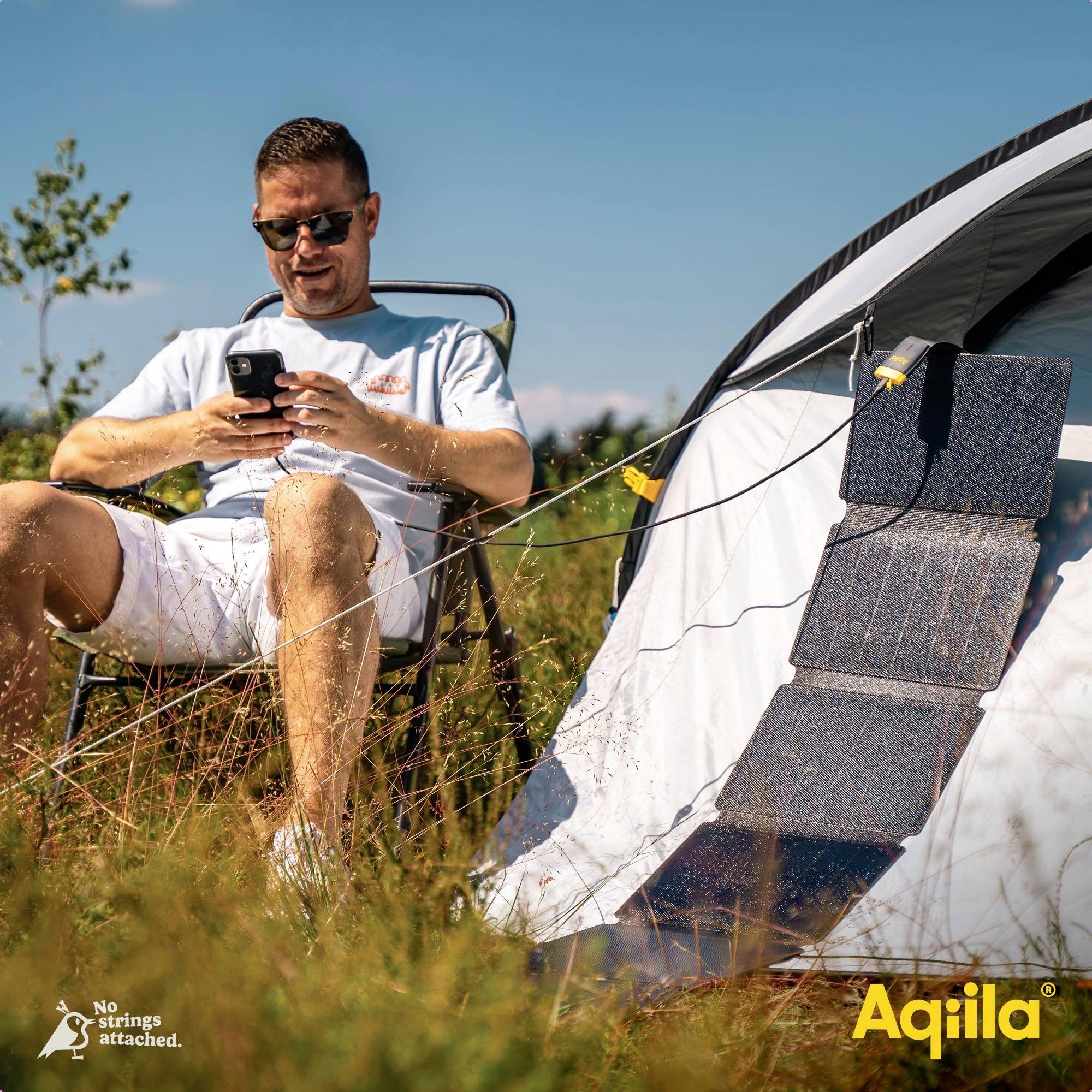 Un homme est assis dans une chaise de camping devant une tente, regardant son téléphone. Un panneau solaire est fixé à la tente, le logo 'Aqiila' est visible.