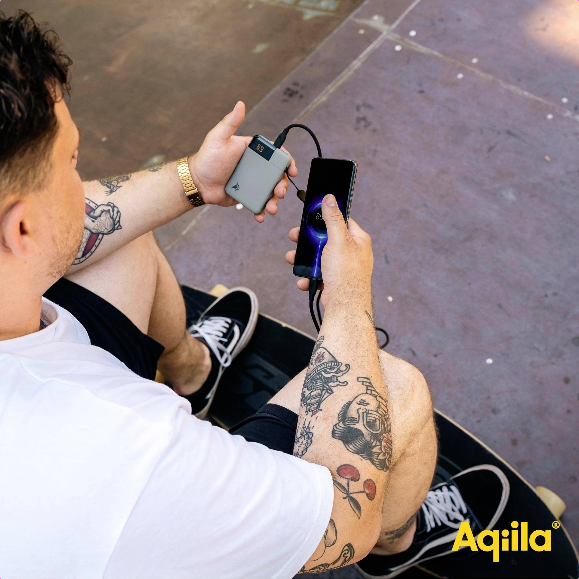 Un homme est assis sur une planche à roulettes, en chargeant son smartphone avec une batterie externe ; la marque 'Aqila' est visible.