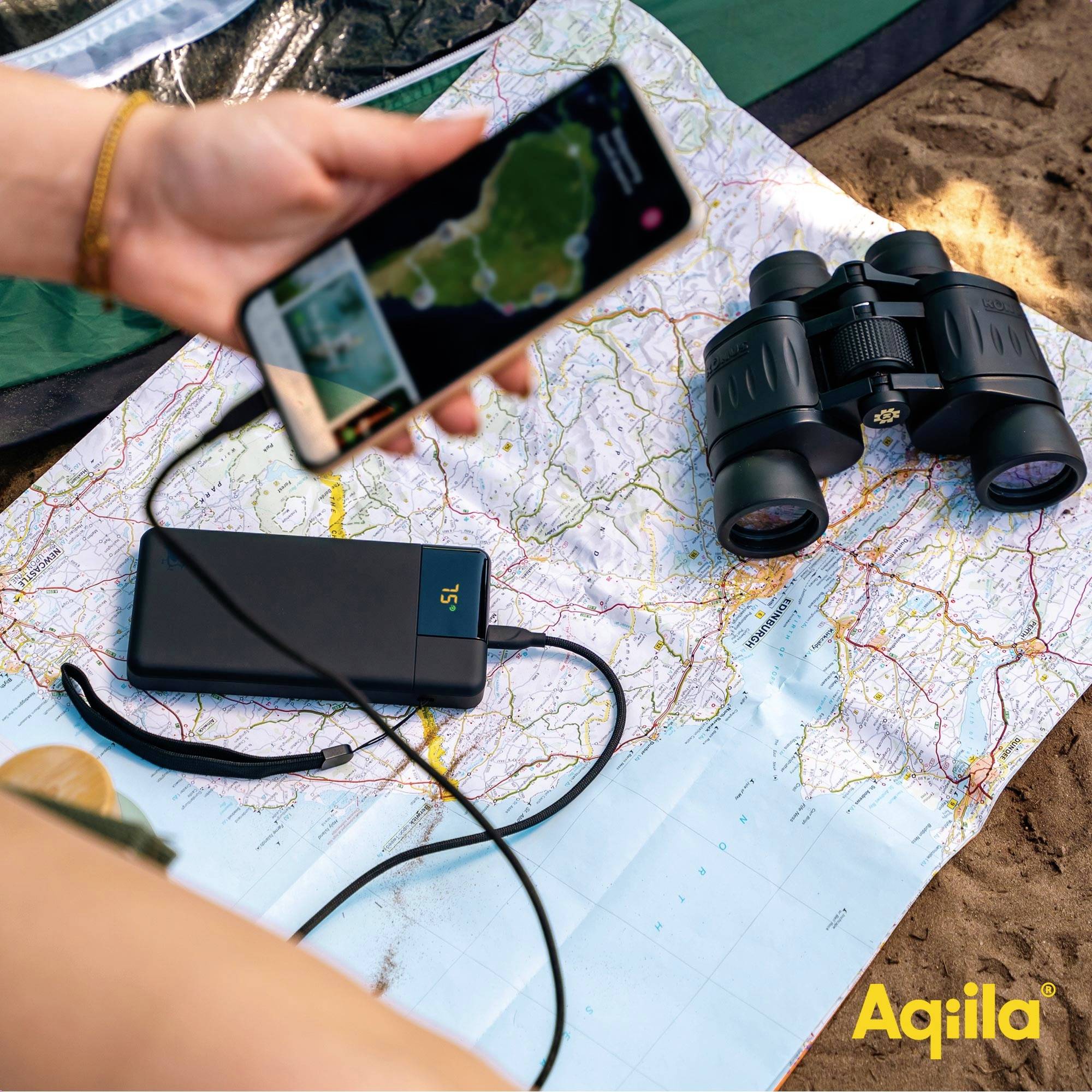Une main tient un smartphone avec une carte, branché à une batterie externe. À côté se trouvent une autre carte et des jumelles.