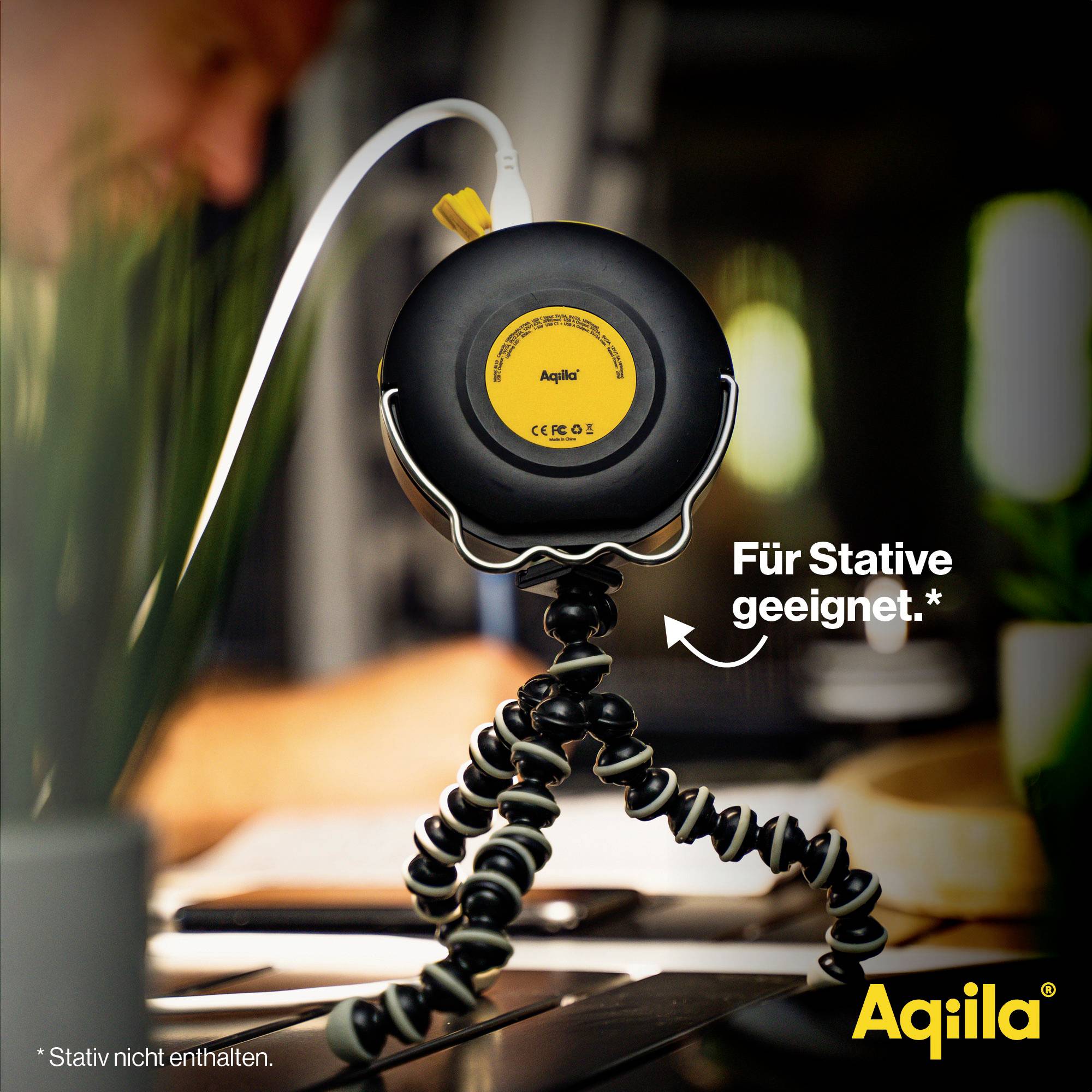 Un haut-parleur portable avec un trépied flexible posé sur une table. Le texte 'Adapté aux trépieds*' est visible. Marque 'Aqila'.