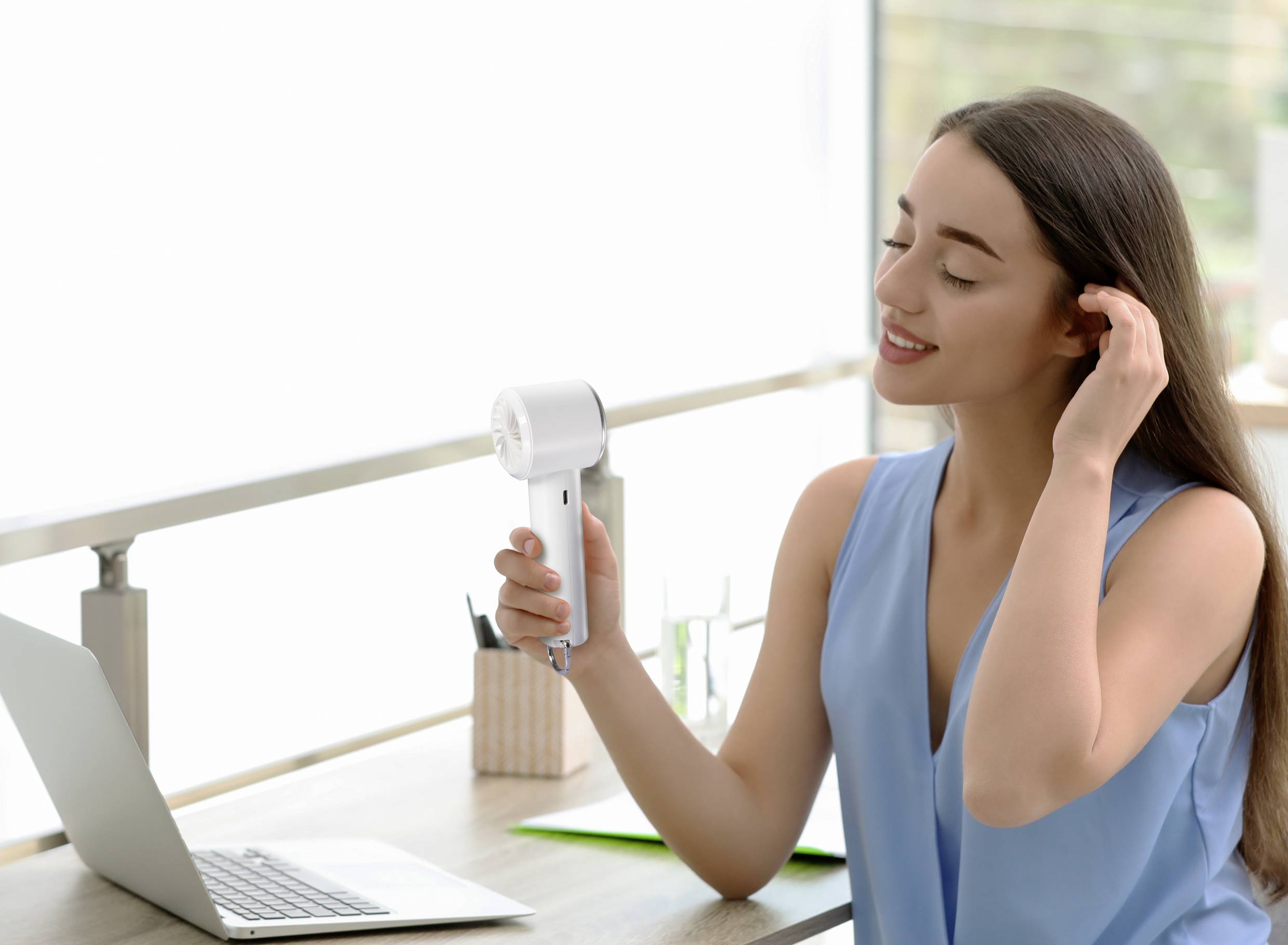 Une femme sèche ses cheveux avec un sèche-cheveux, assise à un bureau avec un ordinateur portable dans une pièce bien éclairée.