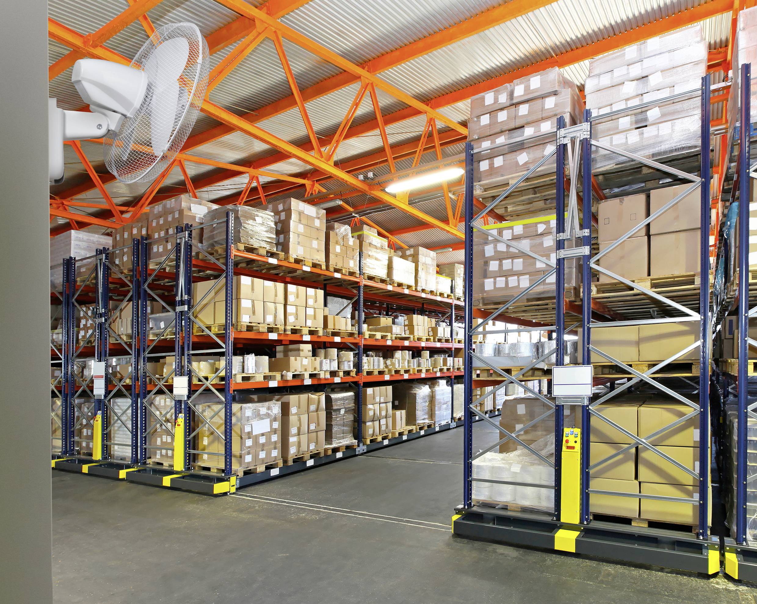 Un entrepôt avec des étagères hautes remplies de cartons et de colis. Un ventilateur sur le mur de gauche assure la ventilation.