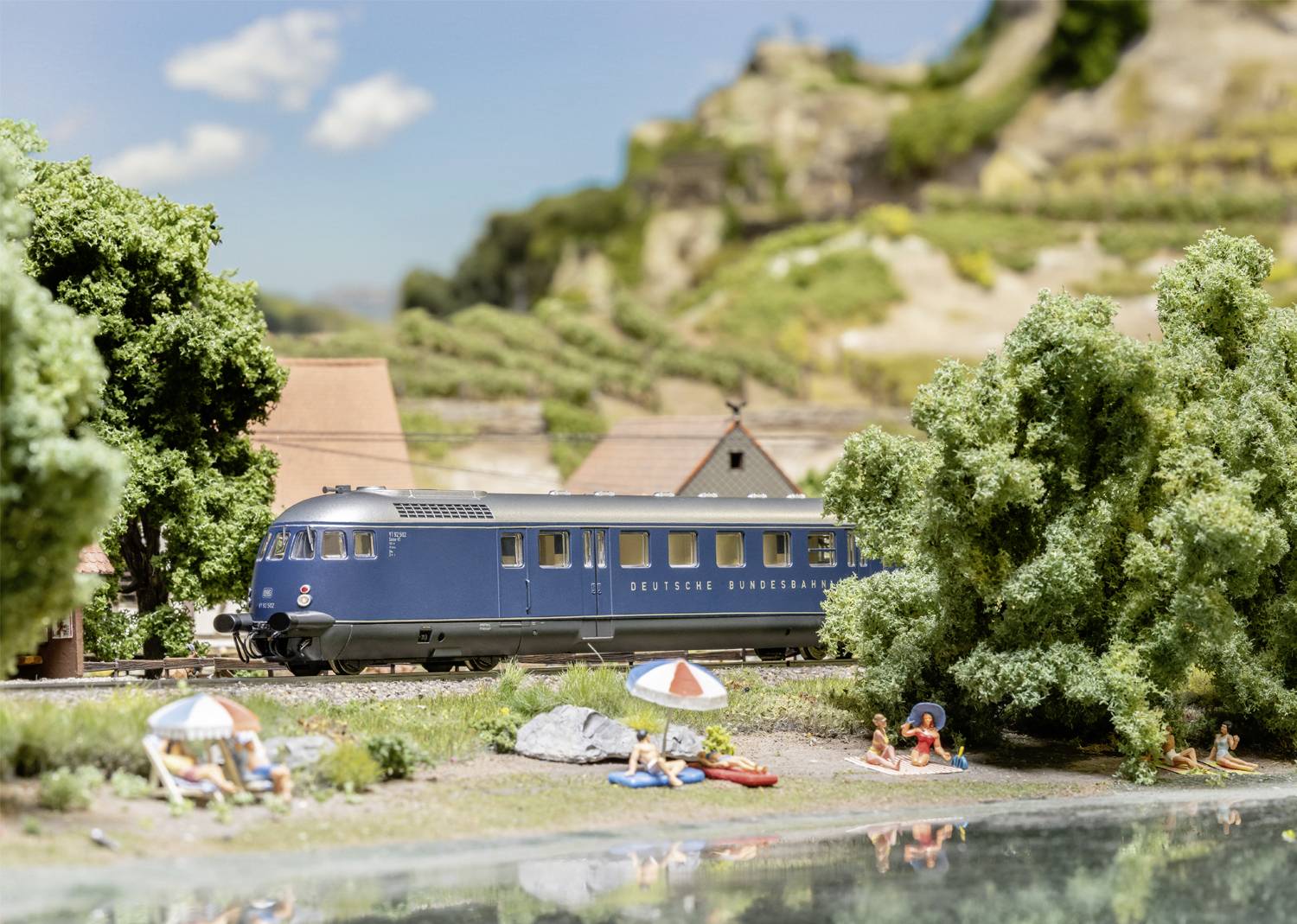 Un train bleu circule par une journée ensoleillée à travers un paysage rural, longeant des arbres verts et des eaux cristallines.