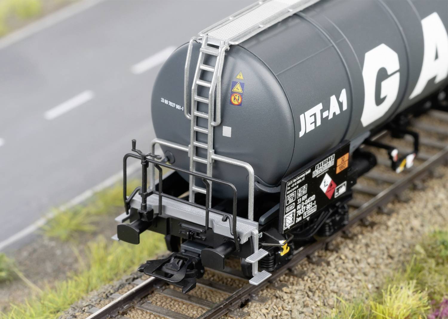 'Modèle d'un train de marchandises avec un wagon-citerne gris, portant l'inscription JET-A1. Il est posé sur des rails, avec une route visible en arrière-plan.'