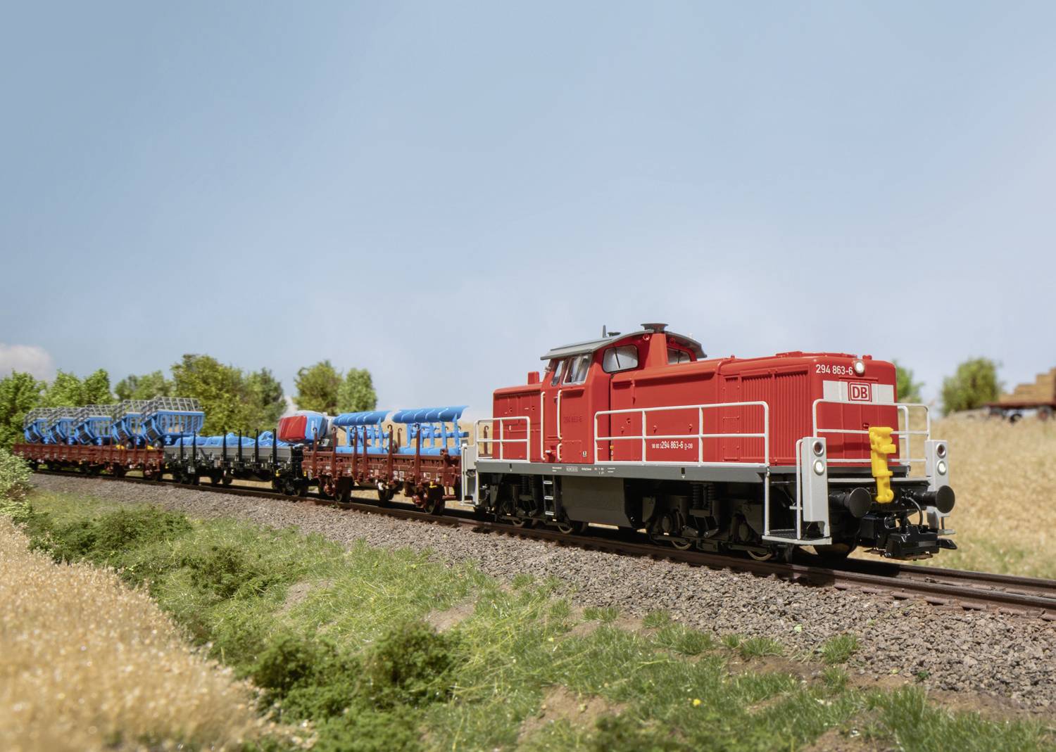 Un train de marchandises rouge tire des wagons chargés à travers un paysage rural par une journée ensoleillée. Des arbres sont visibles en arrière-plan.