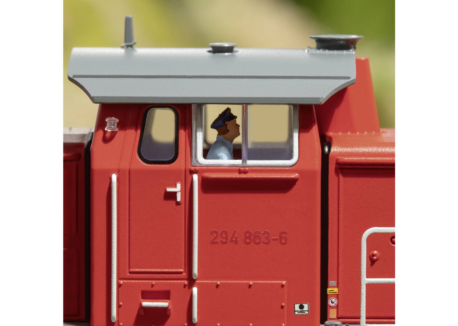 Conducteur de train miniature rouge dans une locomotive portant le numéro '294 863-6', fenêtre visible, fond vert.