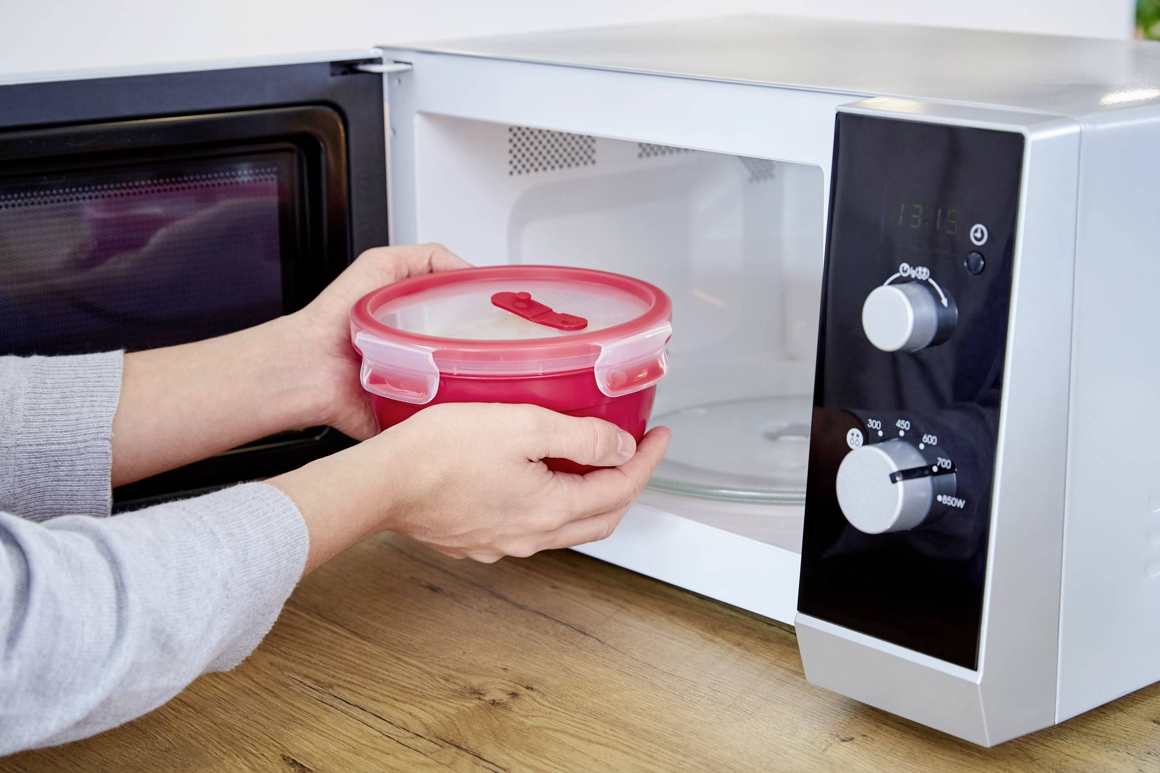 Une personne tient un récipient en plastique rouge et ouvre le micro-ondes pour le placer à l'intérieur. Le micro-ondes est posé sur une surface en bois.