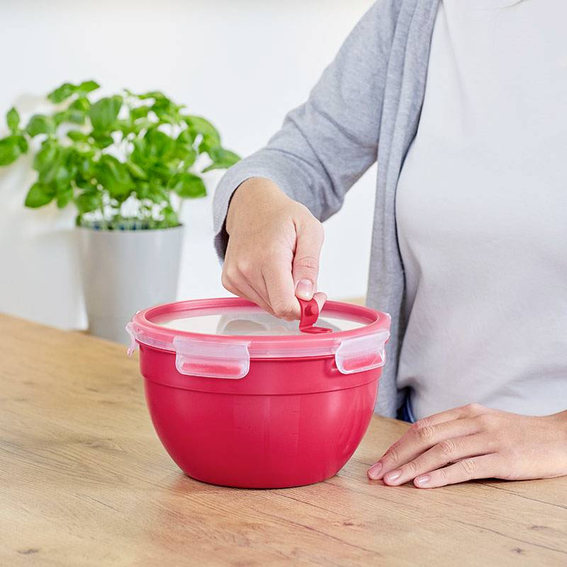 Une personne ouvre le couvercle du bol en plastique rouge d'une seule main sur une table en bois. Une plante est visible en arrière-plan.