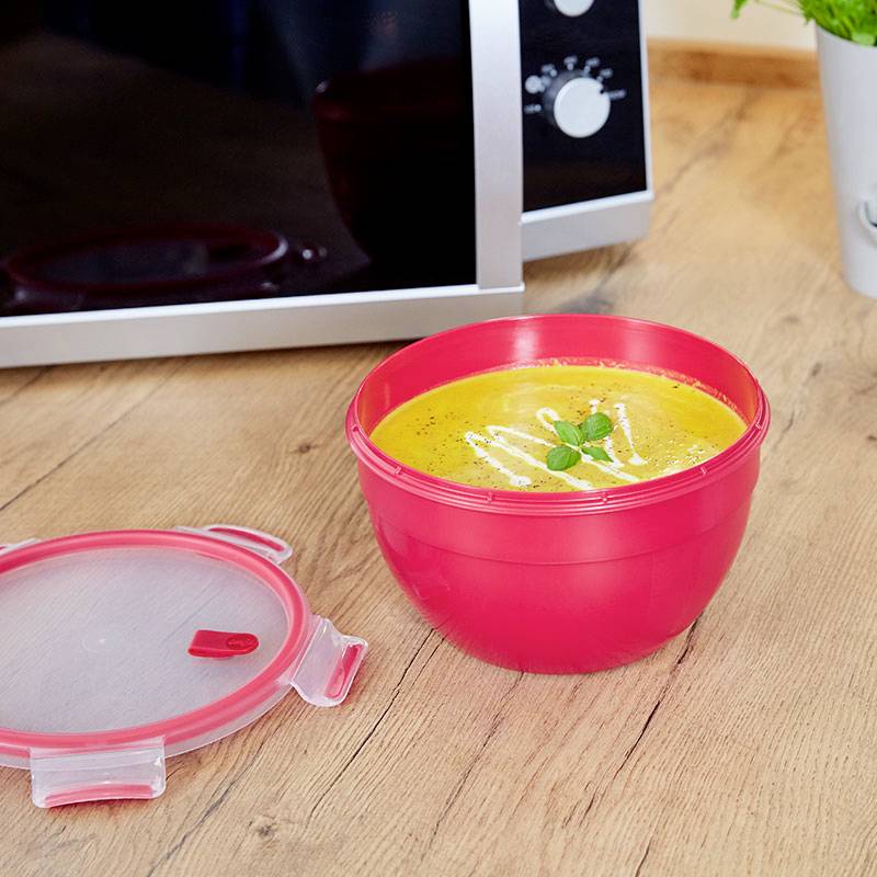 Assiette en plastique rouge remplie de soupe sur une table en bois, à proximité d'un four à micro-ondes. La soupe est garnie d'herbes.