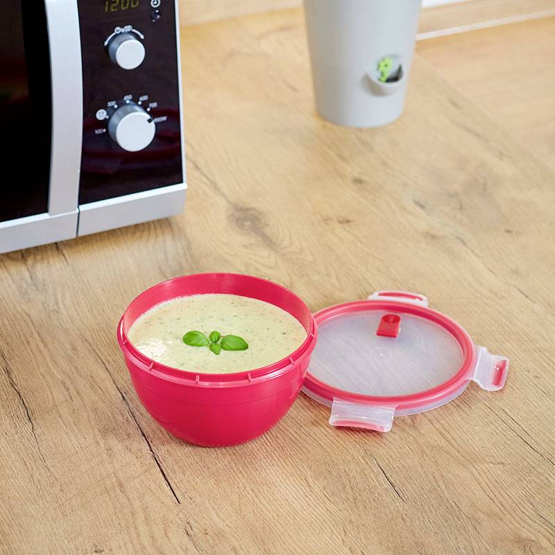 Un bol rouge de soupe sur une table en bois, près d'un four à micro-ondes. Le couvercle du bol est ouvert.