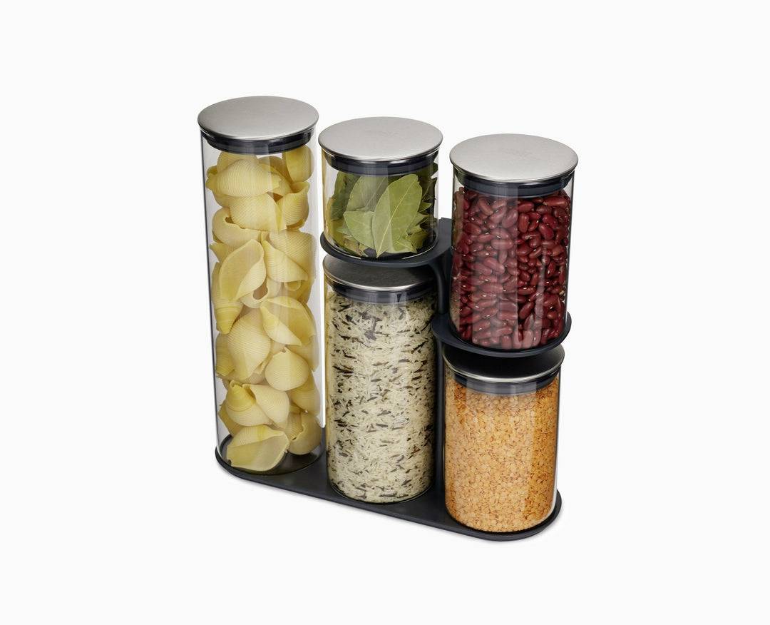 Trois récipients en verre de grande taille et deux de taille moyenne, remplis de pâtes, de feuilles de laurier, de haricots rouges, de riz et de lentilles rouges, sont disposés sur un plateau noir.