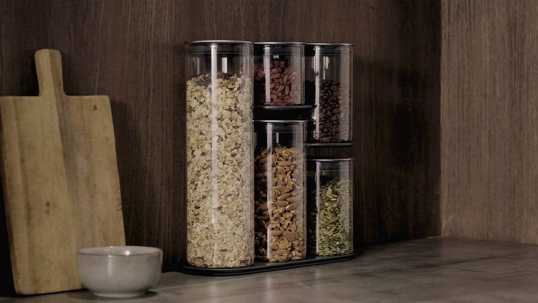 Une petite table en bois présente quatre contenants transparents avec différents aliments comme des céréales et des noix, à côté d'une planche à découper.