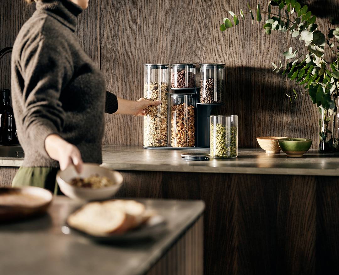 Une personne prépare un muesli dans une cuisine, avec différents contenants de graines et de noix sur le plan de travail.