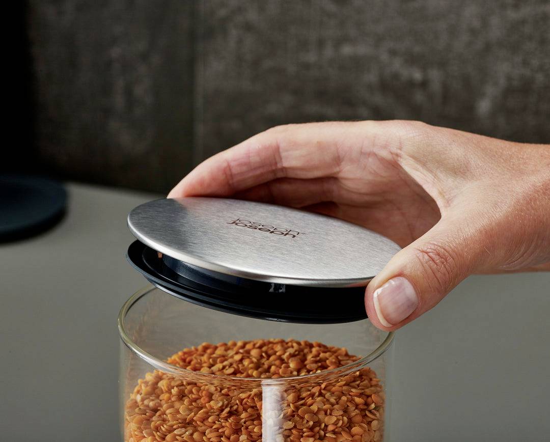 Une main ouvre un bocal à couvercle métallique vissé, rempli de lentilles rouges, sur un plan de travail de cuisine.