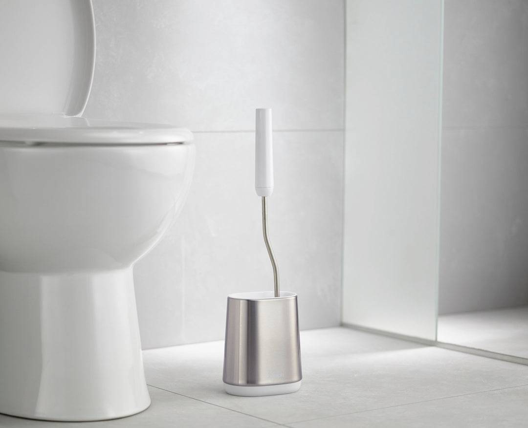 Scène de toilettes avec des toilettes blanches et une brosse de toilette moderne dans un support argenté sur des carreaux clairs.