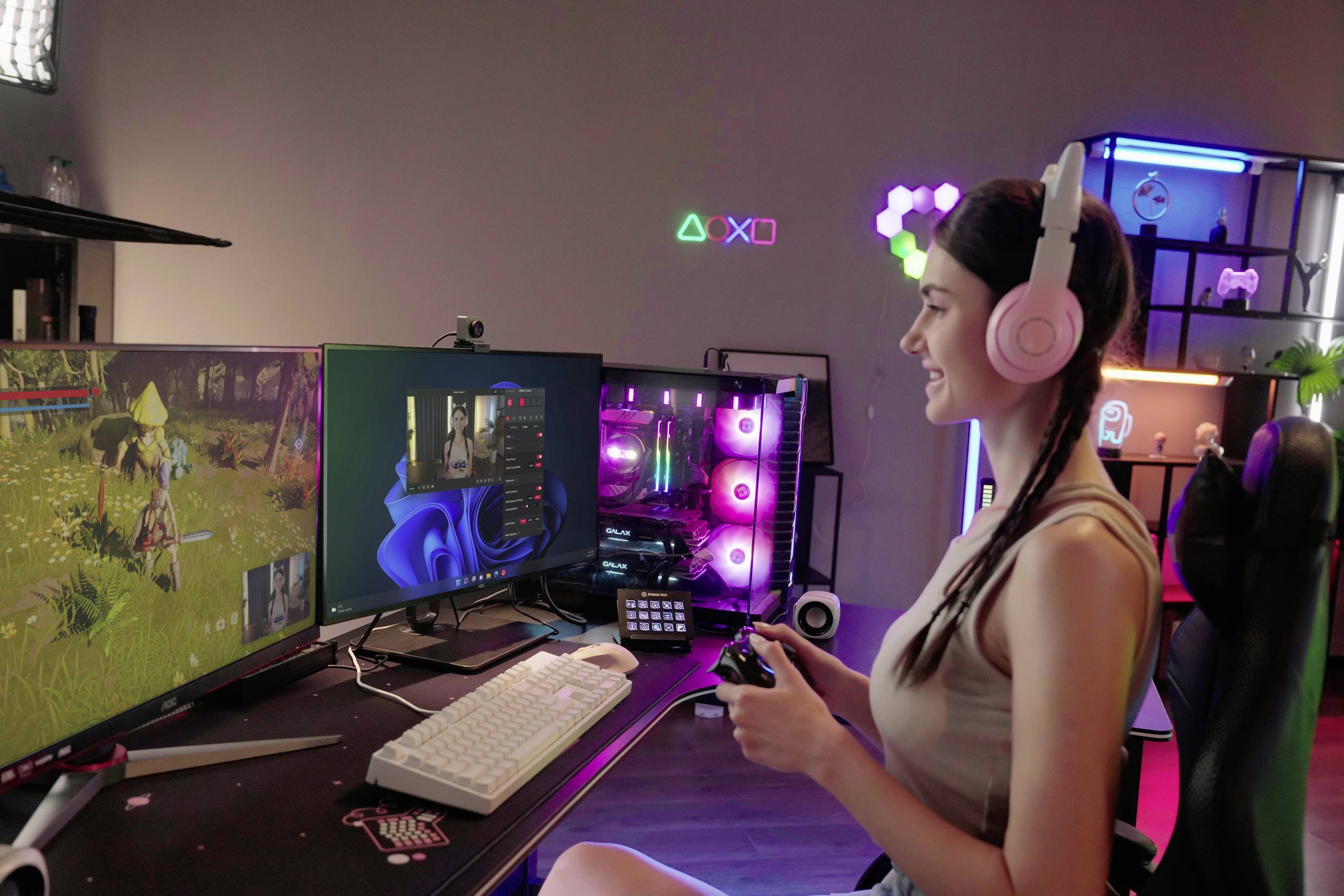 Une femme joue à des jeux vidéo sur un ordinateur à deux écrans. Elle porte des écouteurs et tient une manette de jeu, l'environnement est illuminé de couleurs vives.