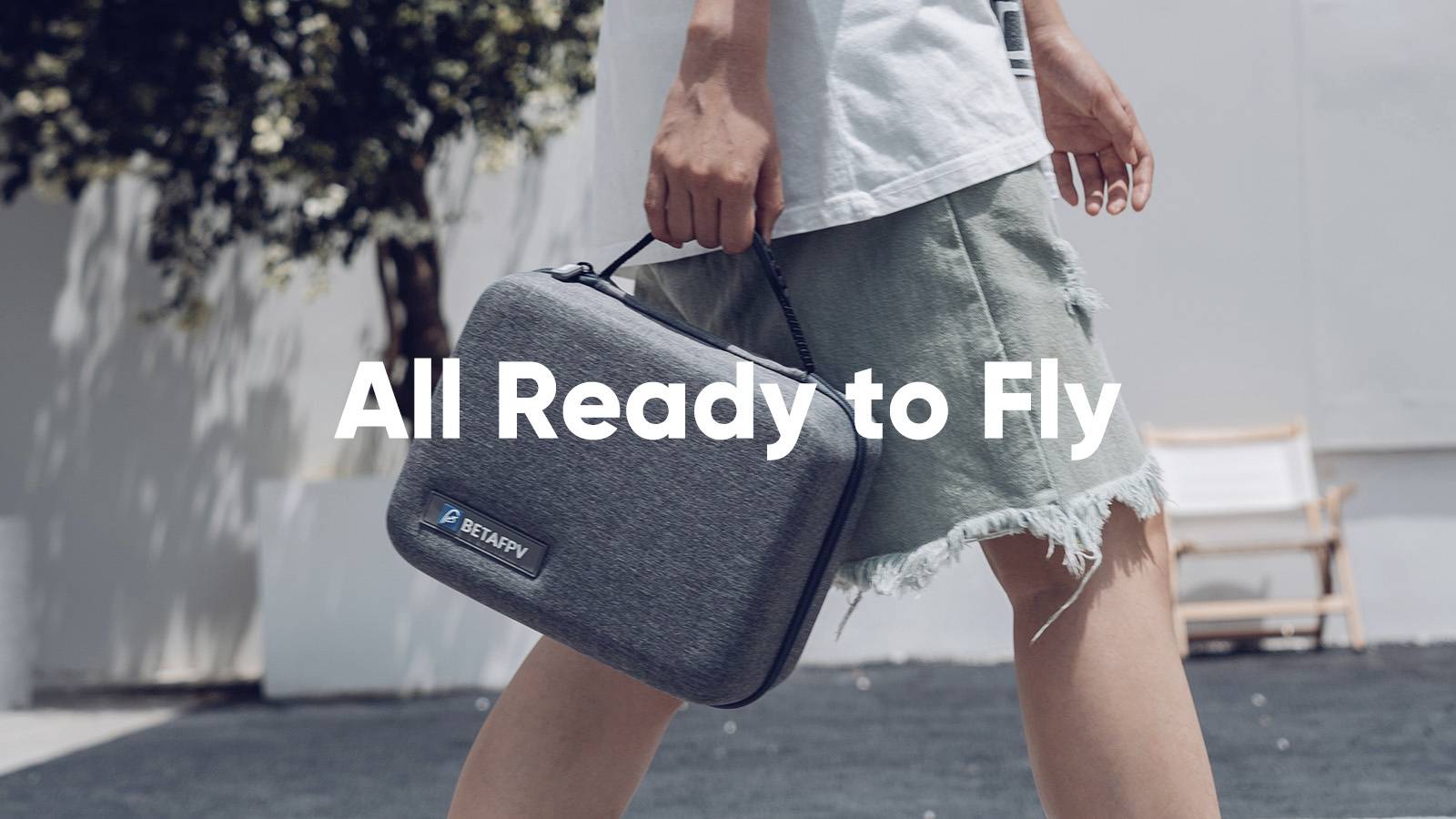 Une personne porte un sac gris sur lequel est inscrit « All Ready to Fly ». La scène semble détendue et estivale.