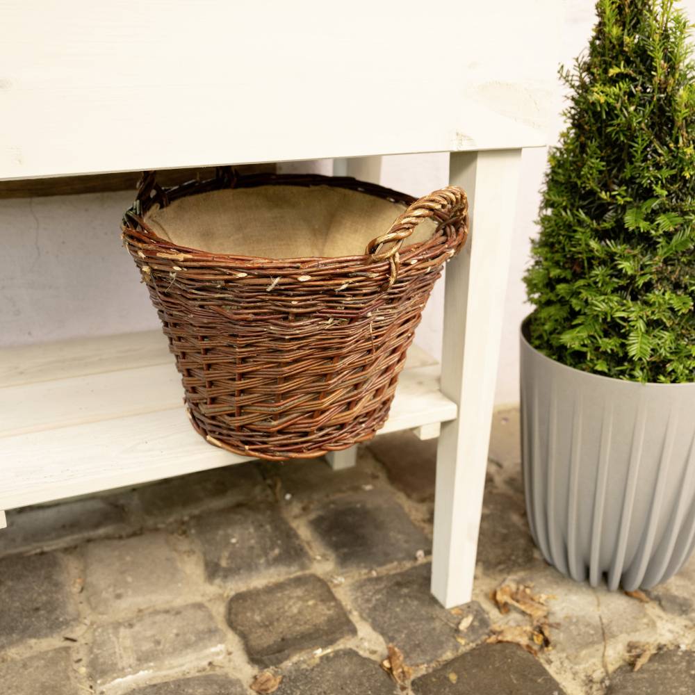 Un panier vide en osier brun est posé sur une étagère blanche à l'extérieur, à côté d'une plante en pot sur un sol pavé.