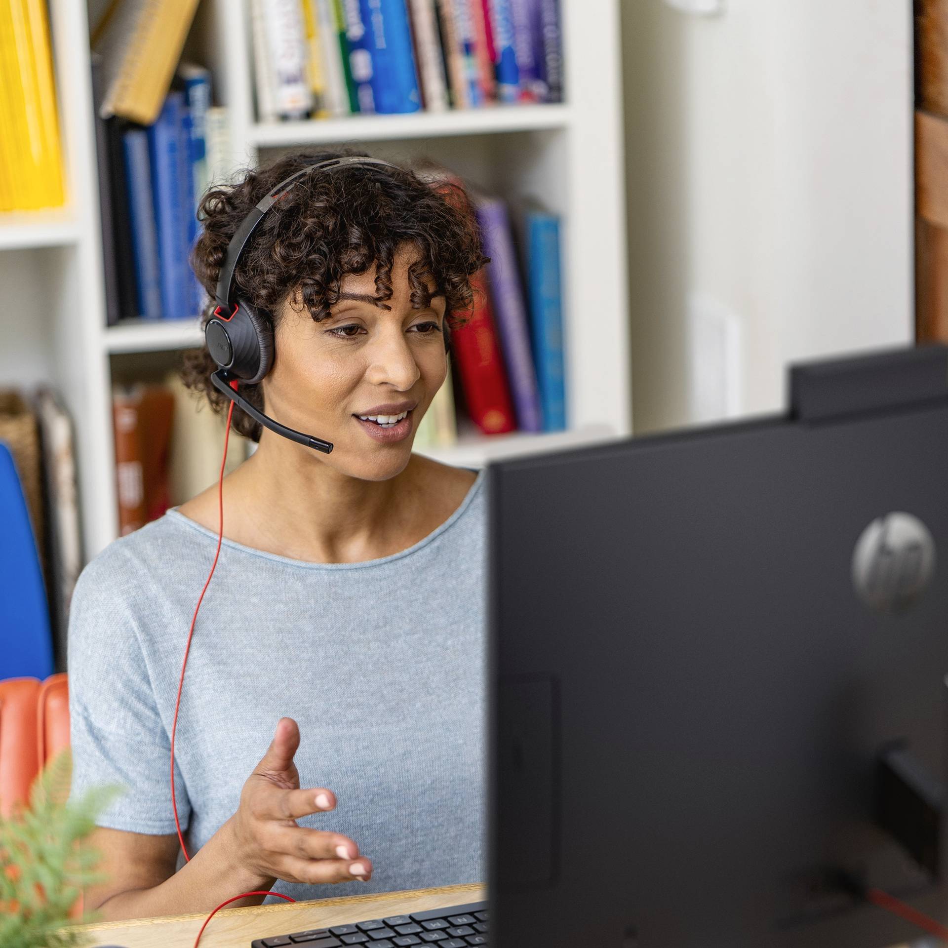 Femme portant des écouteurs et un microphone, souriant lors d'une visioconférence depuis son bureau à domicile. Une bibliothèque est visible en arrière-plan.