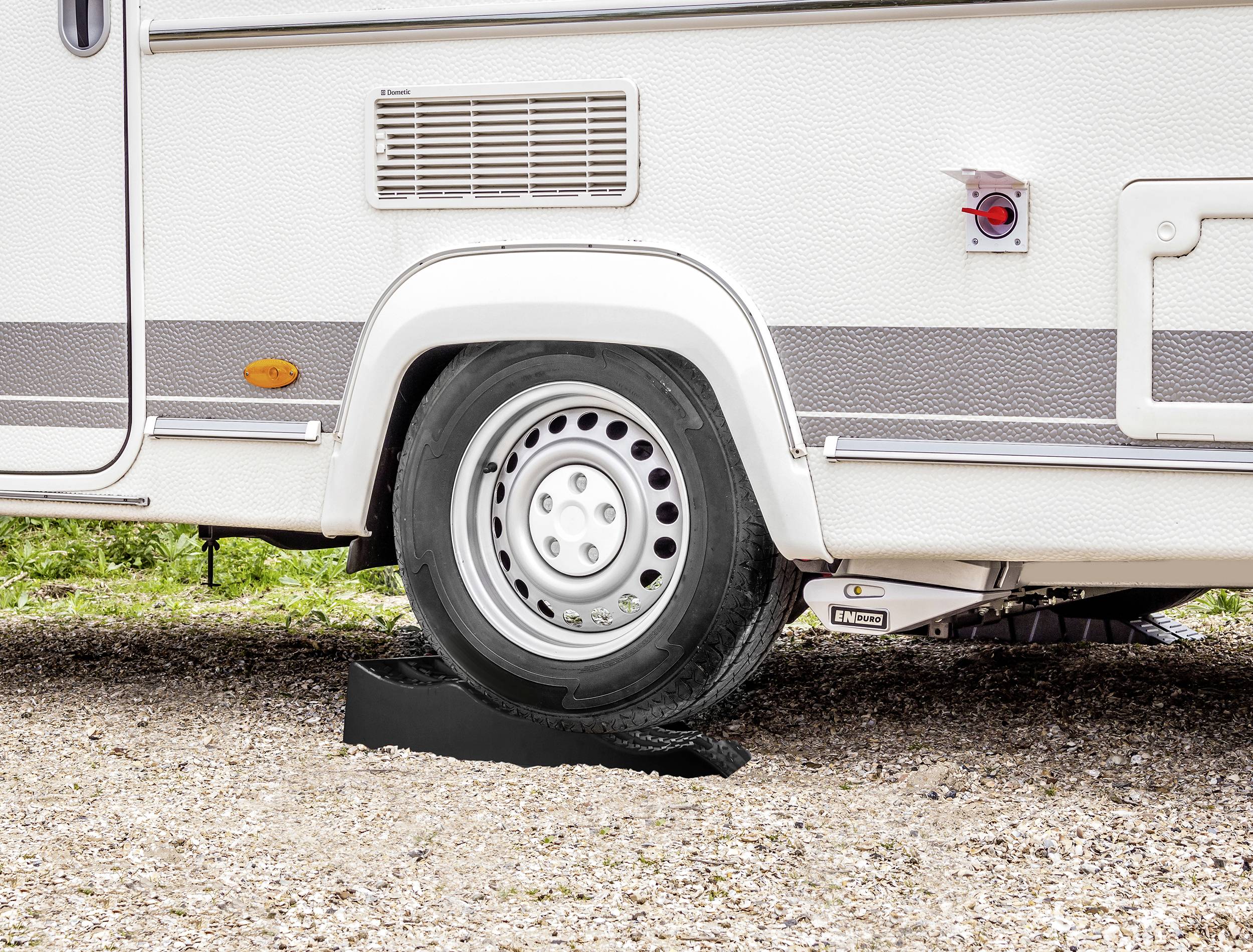 Une caravane est garée sur un sol de gravier, avec le pneu reposant sur un cale-roue noir pour assurer la stabilisation et le nivellement.