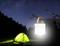 Une tente éclairée dans une forêt la nuit, avec une lanterne lumineuse accrochée à une branche au premier plan. Des étoiles dans le ciel.