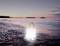 Une lanterne LED brillante se dresse sur la rive d'une plage au coucher du soleil, se reflétant dans l'eau. En arrière-plan : la mer et des îles dans la pénombre.