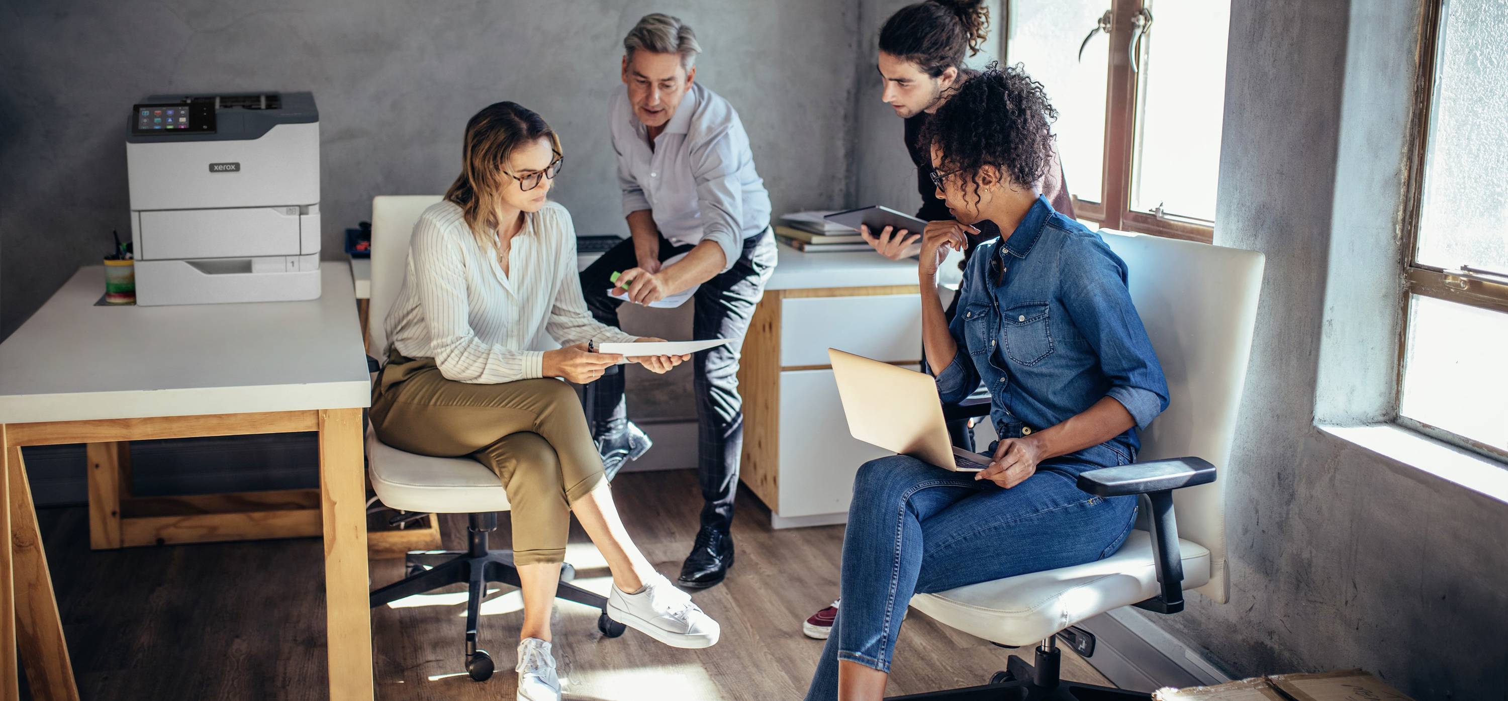 Quatre personnes discutent de documents dans un bureau. Une imprimante est posée sur une table en arrière-plan. Une lumière naturelle entre dans la pièce.