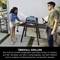 Une femme apporte des légumes à un barbecue, un homme tient une boisson. Le barbecue est installé dans le jardin à côté d'une table avec des ustensiles.