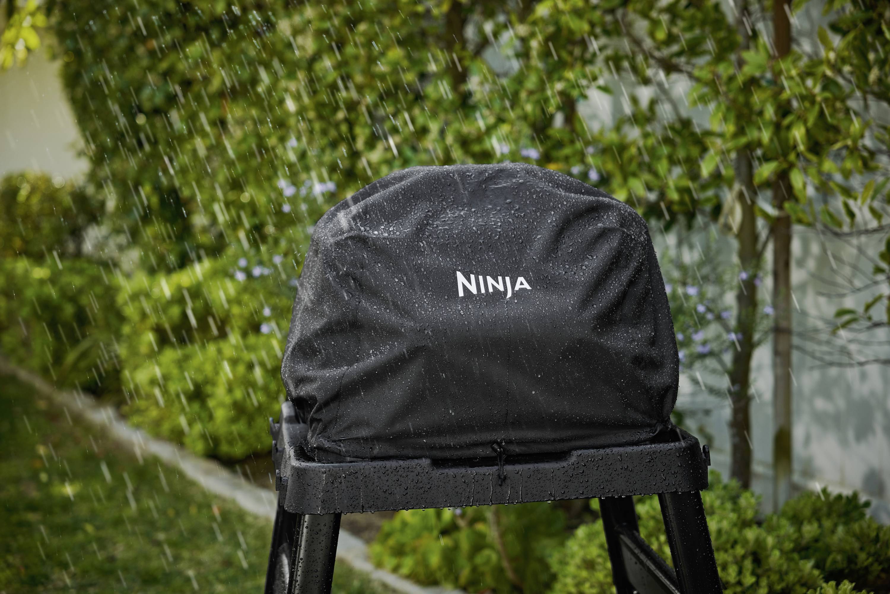 Un appareil couvert d'une bâche noire sous la pluie, posé sur un support dans le jardin. La bâche porte l'inscription 'Ninja'.