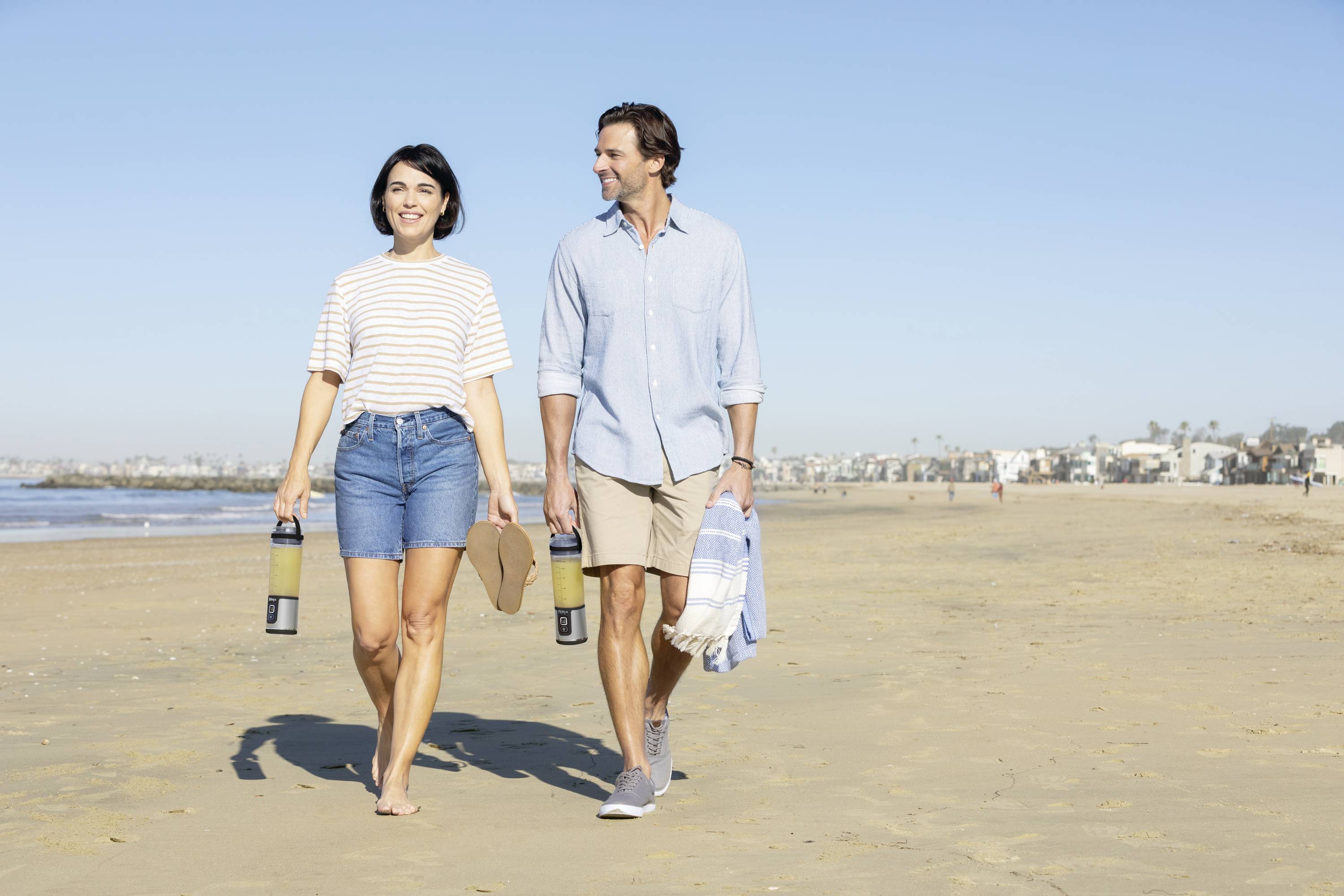 Un couple se promène en souriant le long de la plage, portant des sacs de plage et tenant leurs chaussures à la main. Des maisons et la mer sont visibles en arrière-plan.