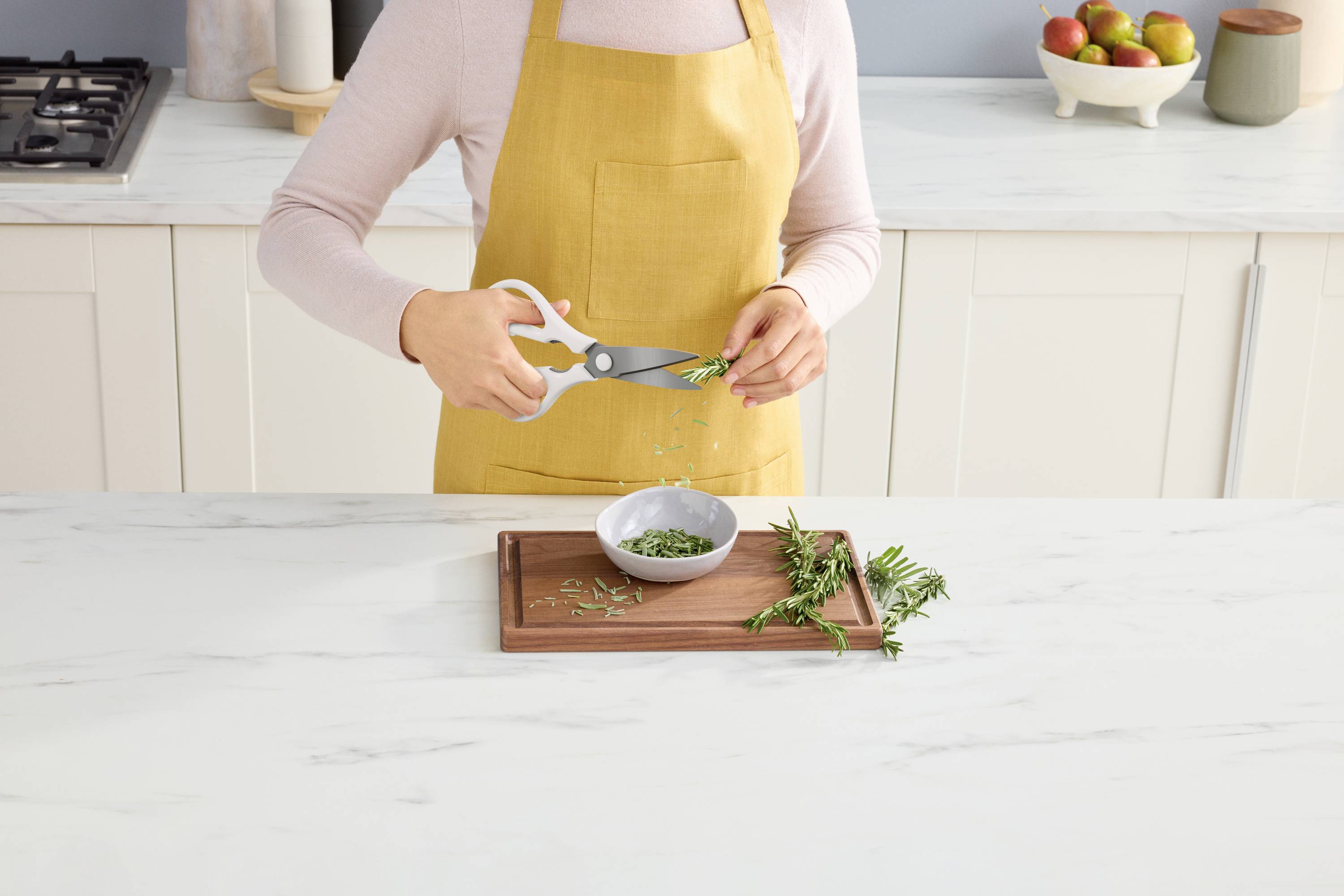 Une personne portant un tablier jaune coupe des herbes fraîches avec des ciseaux au-dessus d'un bol dans une cuisine moderne.
