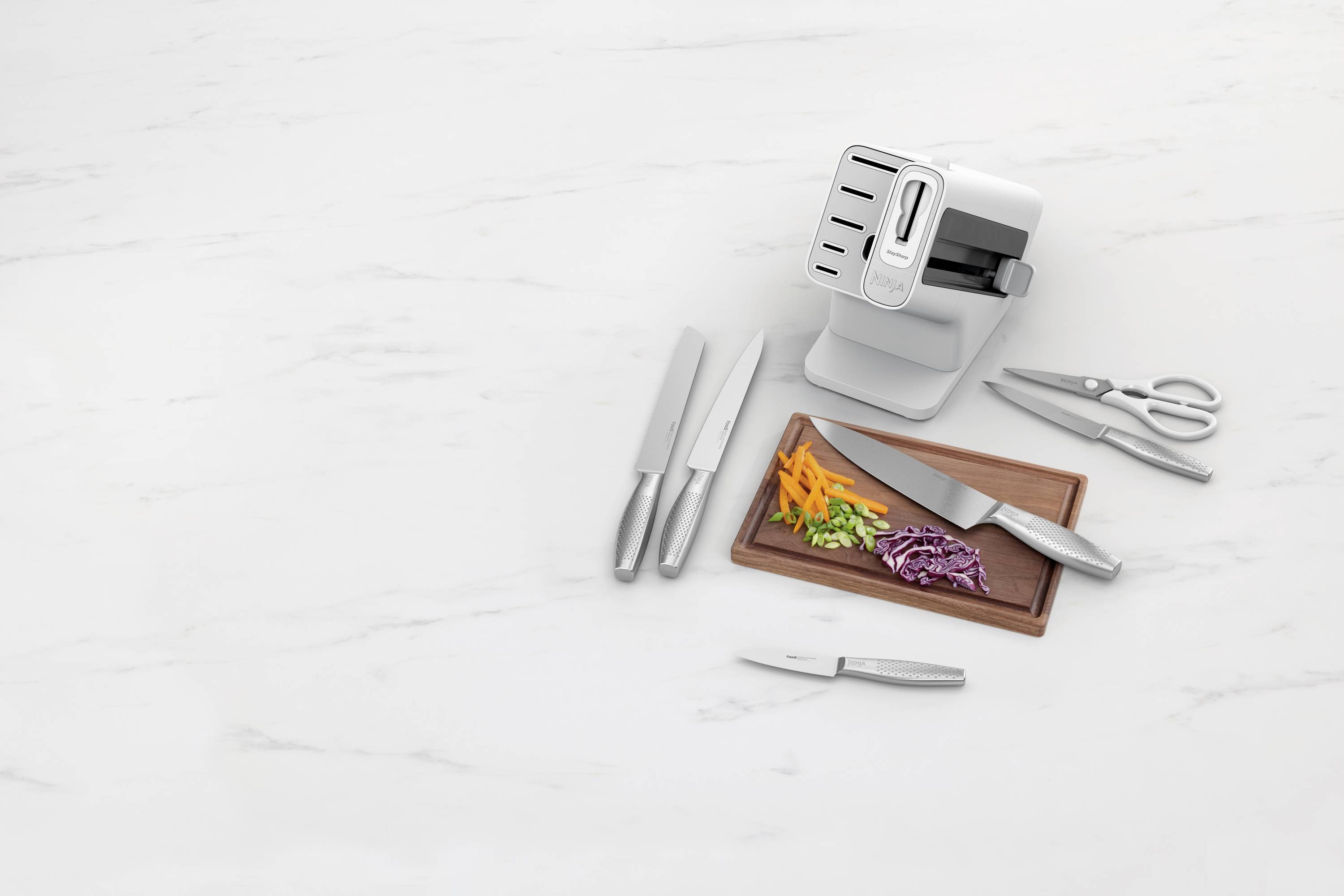 Un bloc de couteaux contenant plusieurs couteaux, une paire de ciseaux et des légumes coupés sur une planche, placé sur une surface en marbre.