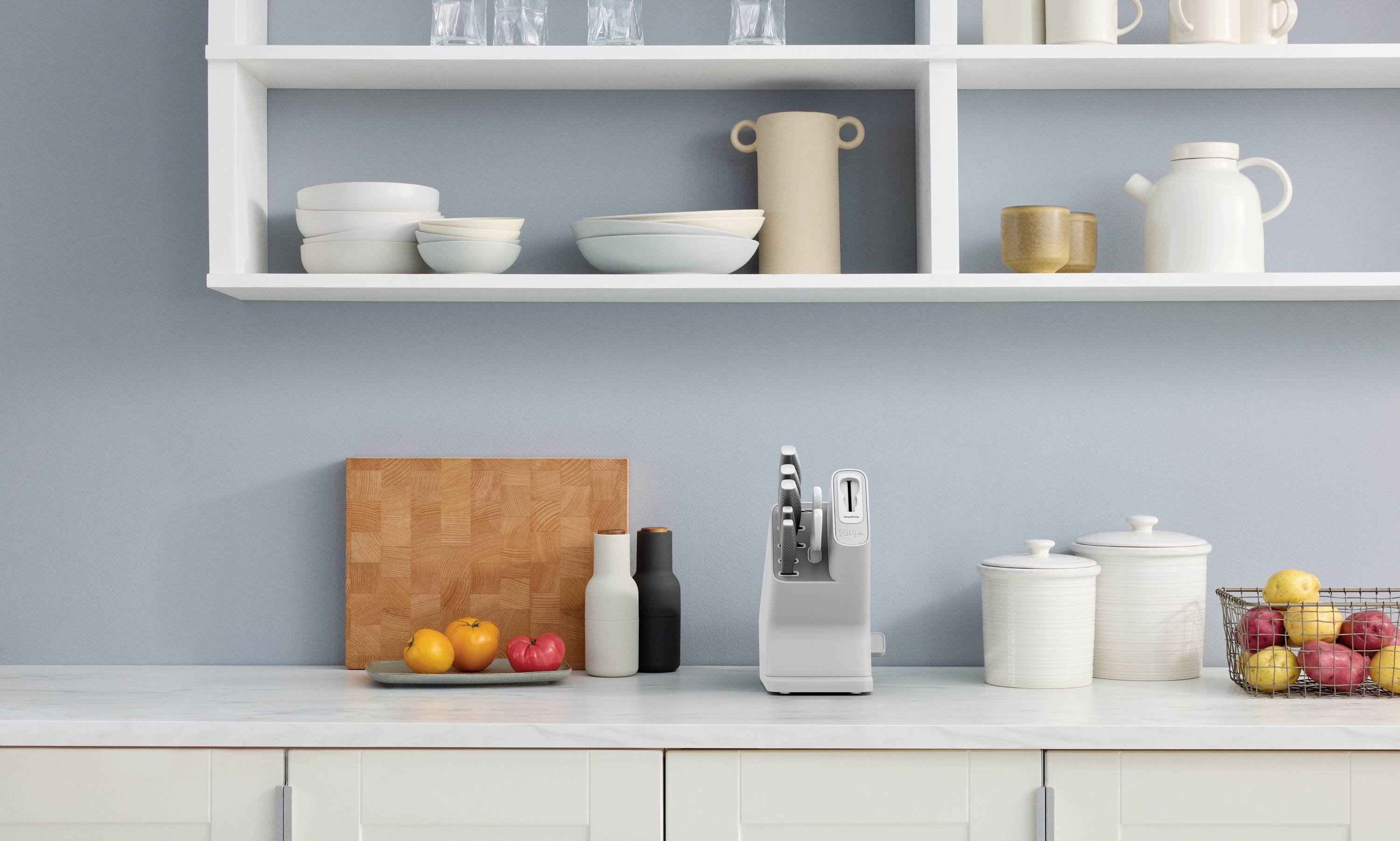 Une cuisine moderne avec des étagères blanches et un plan de travail. Un panier de fruits et un bloc de couteaux sur le plan de travail, des pièces de vaisselle décoratives sur l'étagère.