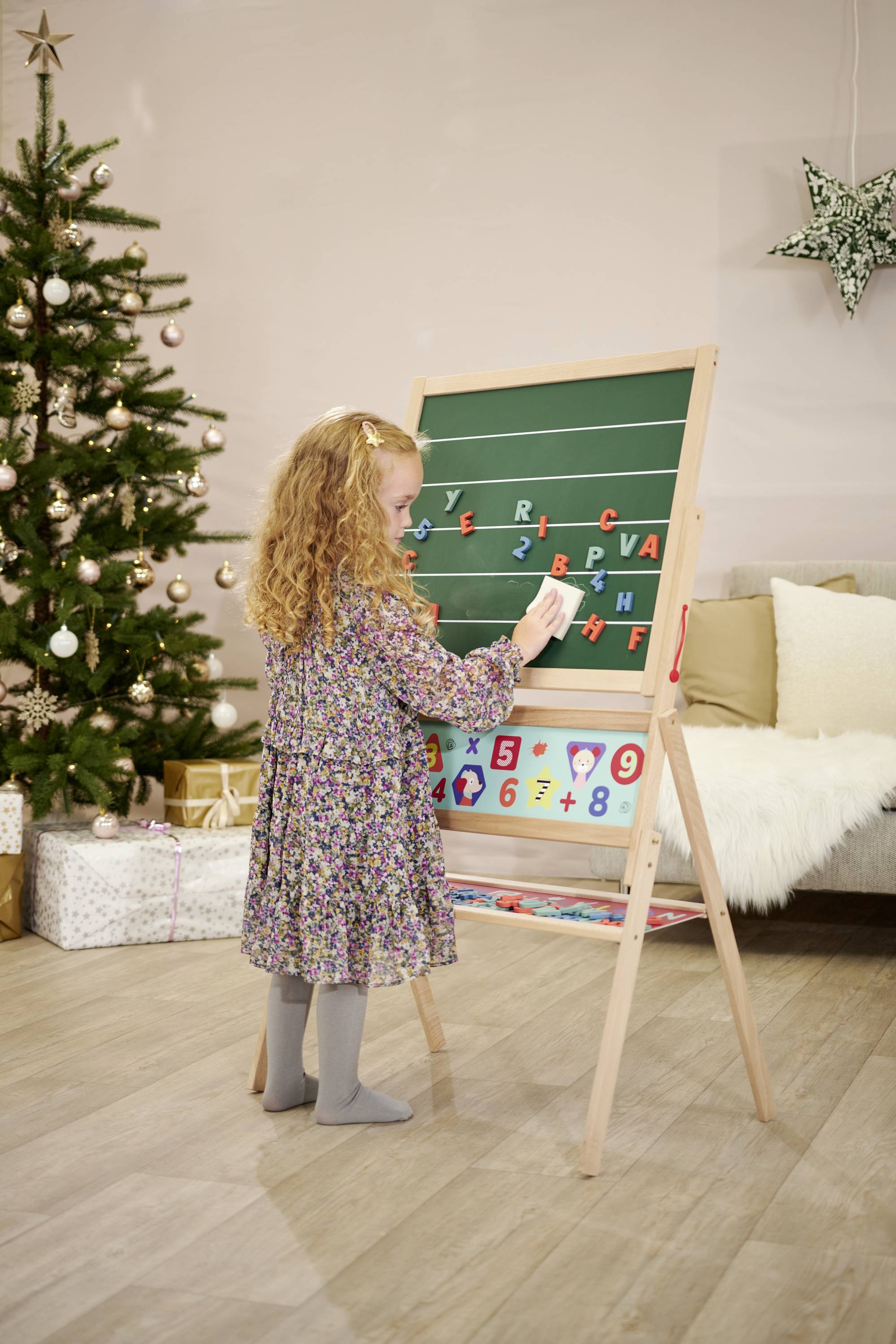 Un enfant écrit des lettres sur un tableau vert à côté d'un arbre de Noël décoré. Des cadeaux sont disposés en arrière-plan.