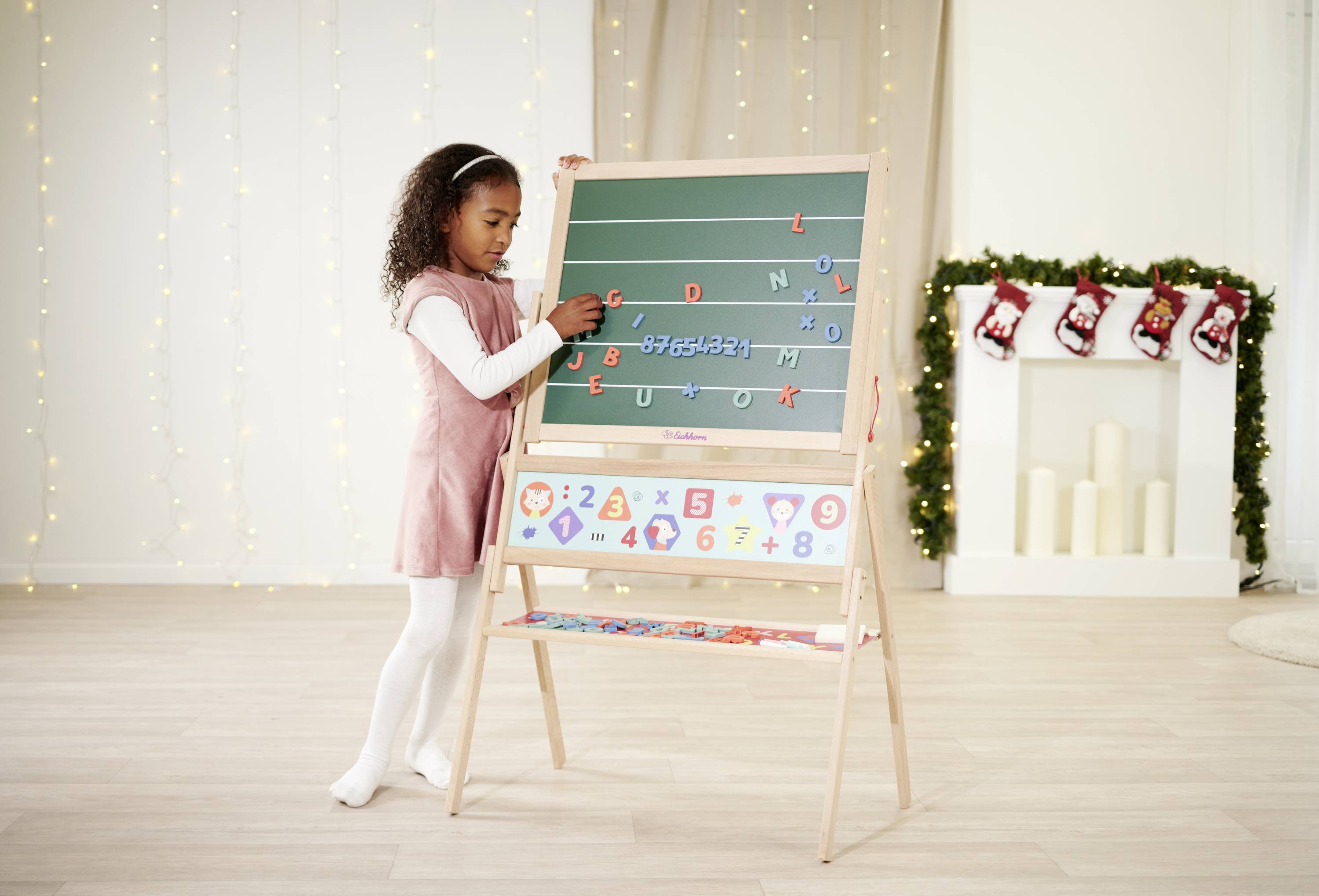 Un enfant joue avec des lettres magnétiques sur un tableau dans une pièce décorée pour Noël. Des guirlandes lumineuses et des chaussettes sont visibles en arrière-plan.