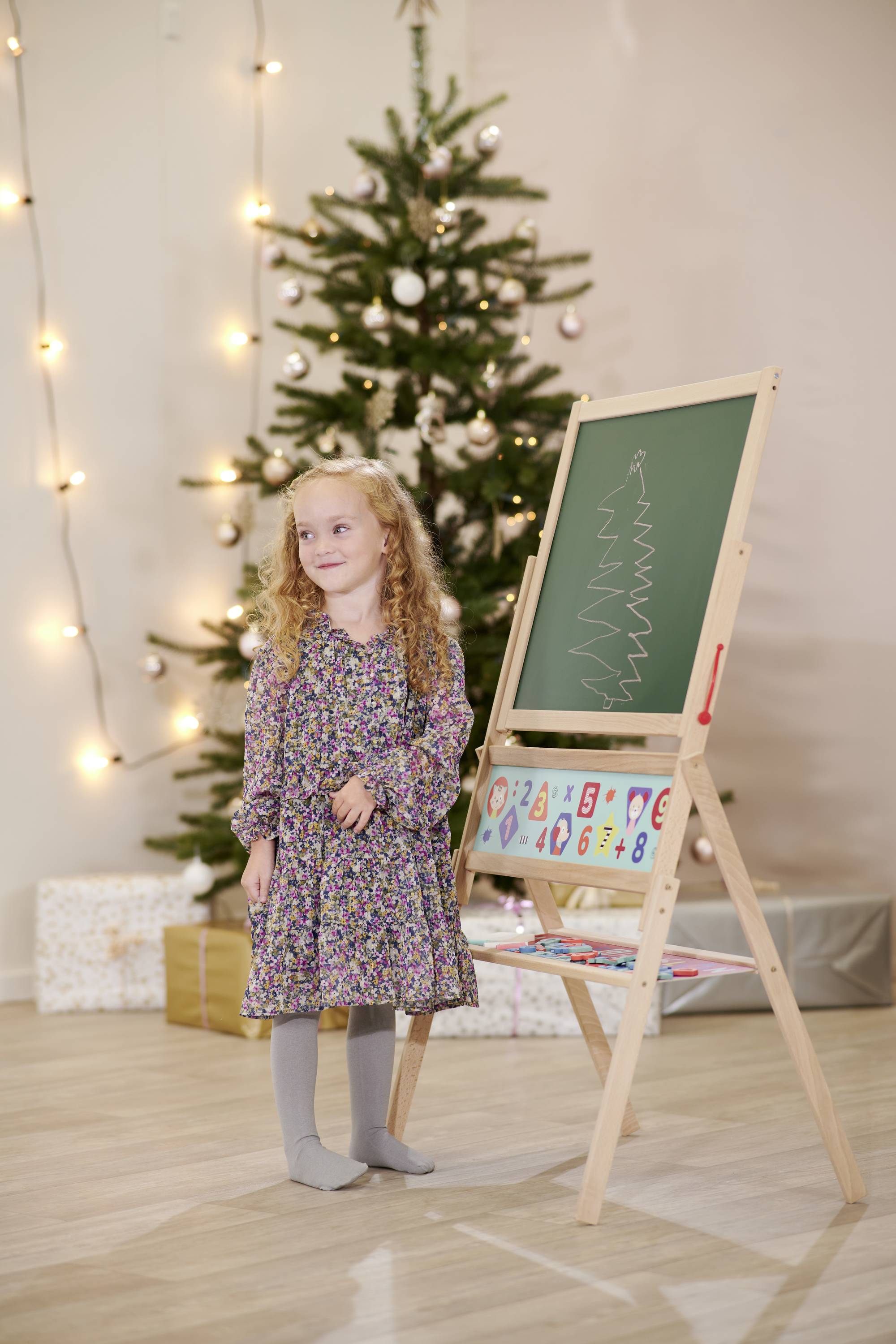 Une jeune fille se tient à côté d'un tableau sur lequel un sapin de Noël est dessiné. En arrière-plan, un sapin de Noël décoré brille.