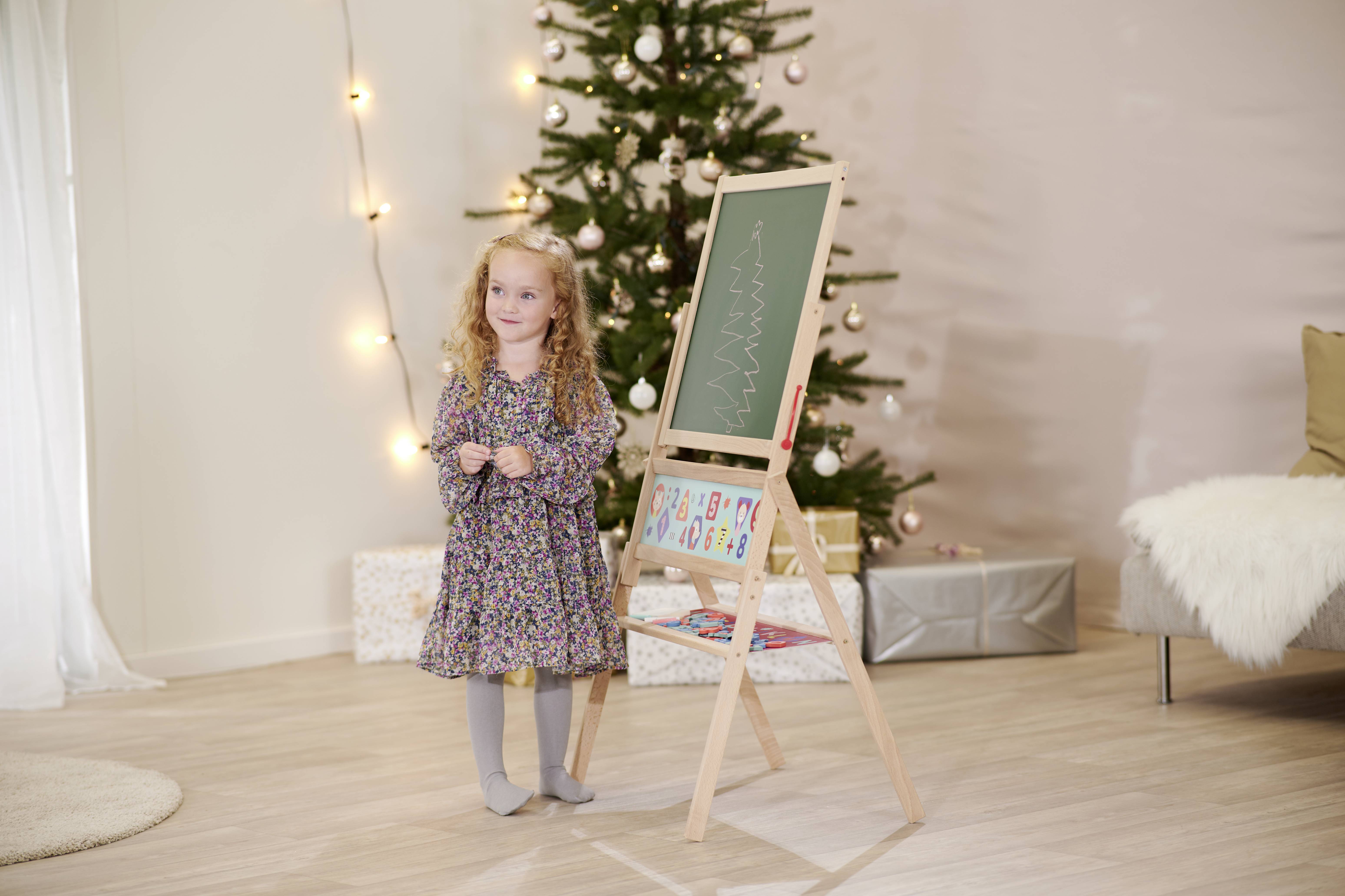 Une petite fille se tient à côté d'un tableau sur lequel est dessiné un arbre de Noël. En arrière-plan, un sapin de Noël décoré est visible.