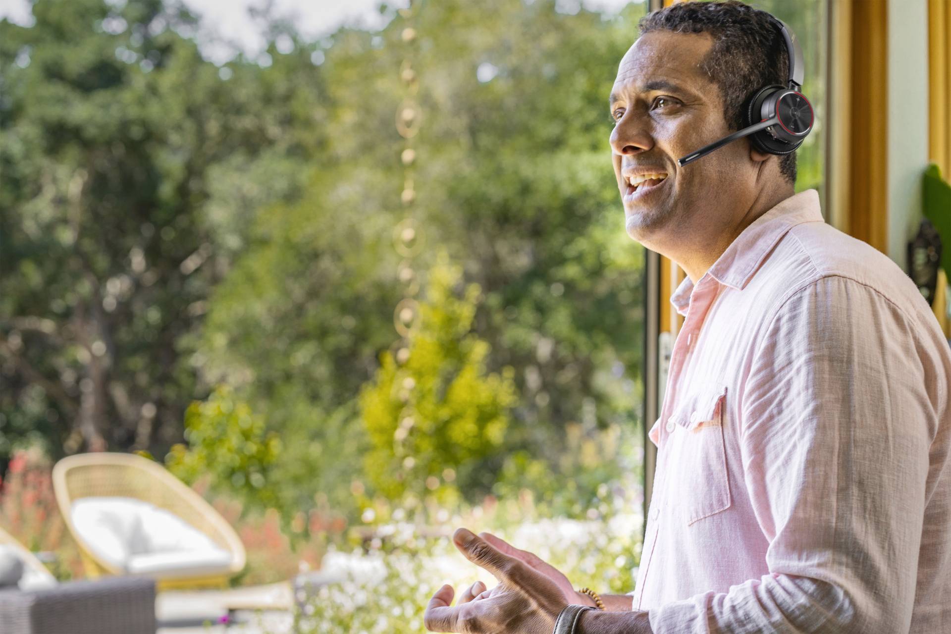 Un homme portant un casque sourit tout en parlant, debout devant une fenêtre donnant sur un jardin ensoleillé.