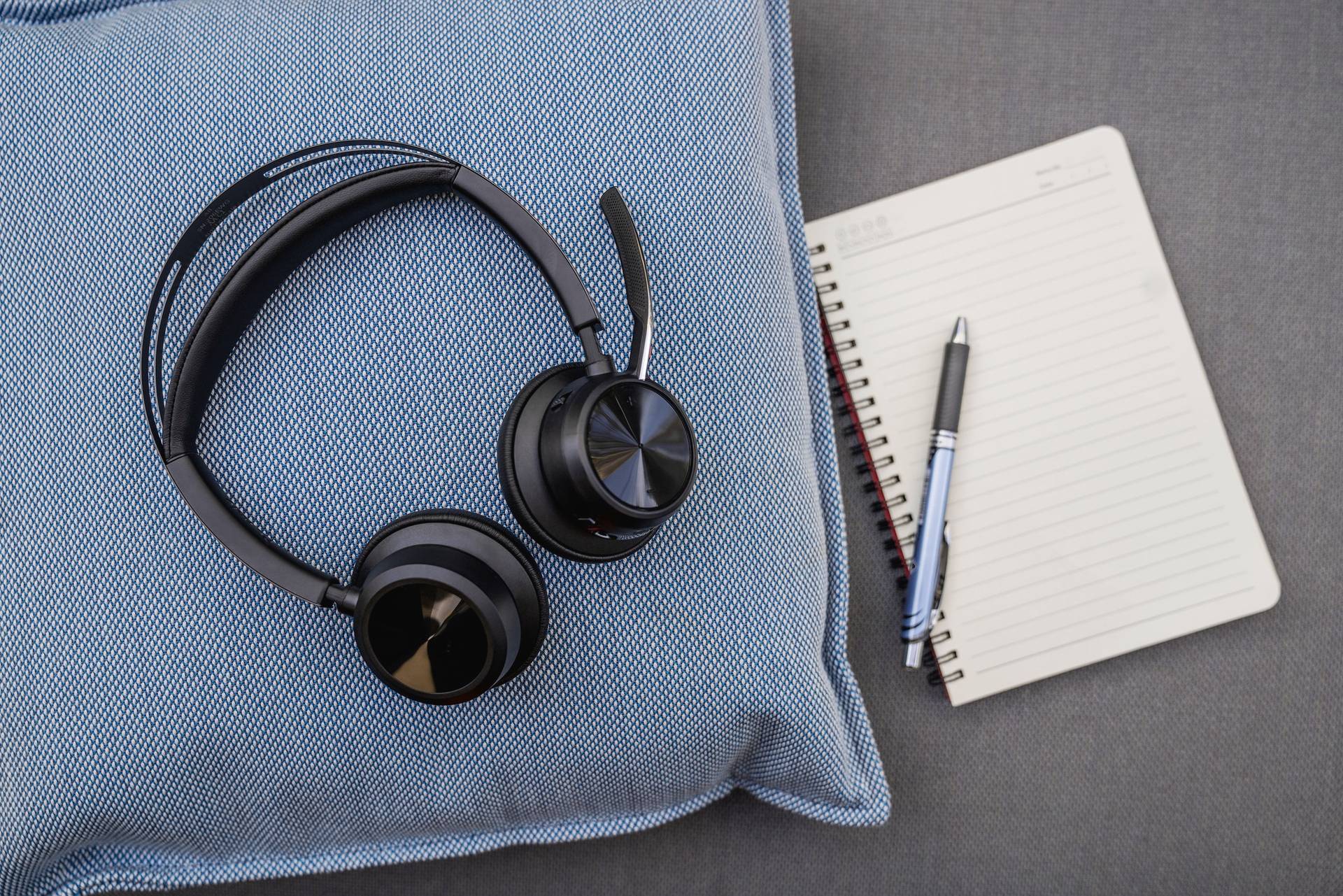 Écouteurs noirs sur un coussin bleu ; à côté, un carnet ouvert avec un stylo à bille sur un fond gris.