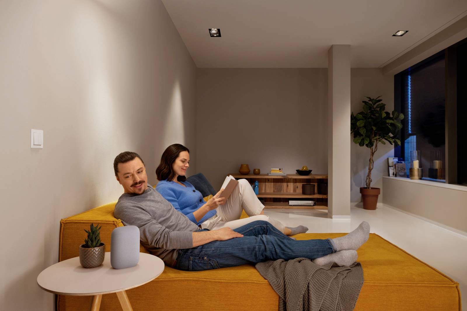 Un homme et une femme se détendent sur un canapé dans un salon au design moderne. Tous deux lisent un livre ou regardent une tablette.