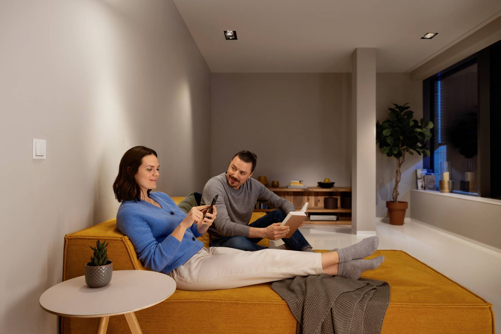 Une femme et un homme se détendent sur un canapé jaune. La femme regarde son téléphone portable, tandis que l'homme fixe une tablette.