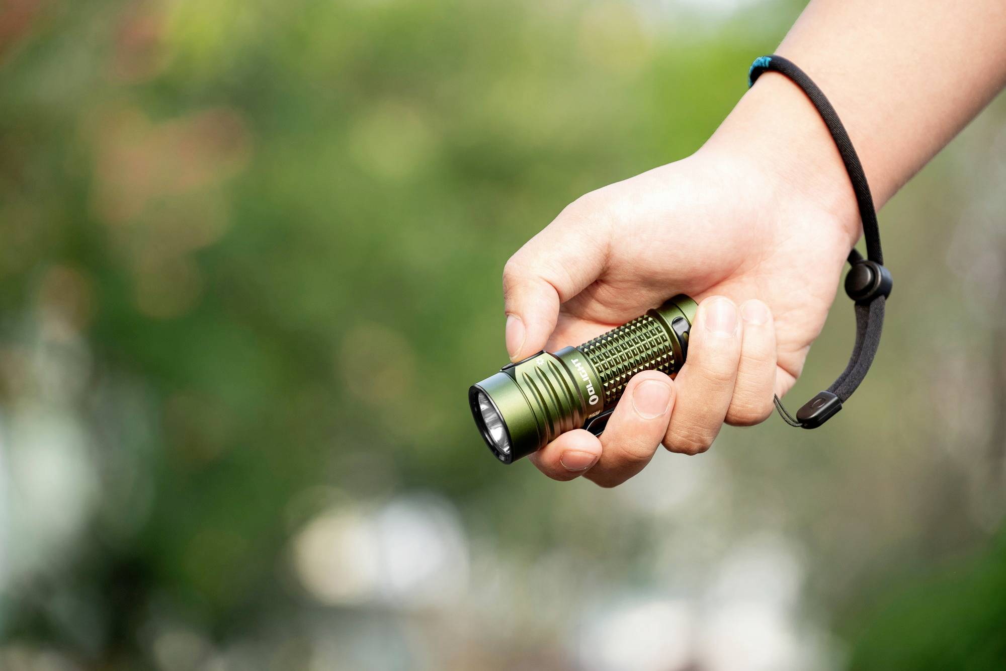 Une main tient une petite lampe de poche verte à l'extérieur. Des arbres flous sont visibles en arrière-plan.
