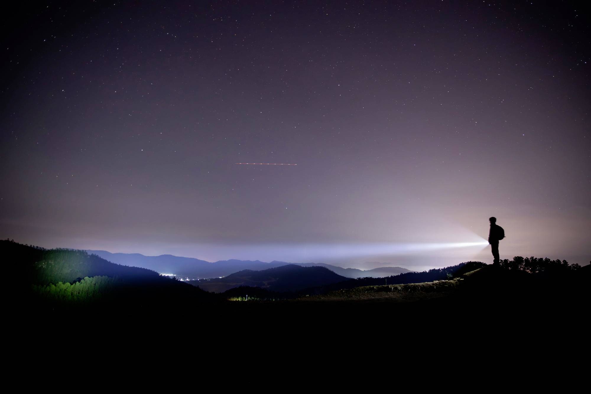 Une personne se tient sur une colline dans la nuit, regardant au loin et tenant une lampe de poche qui projette un faisceau lumineux.