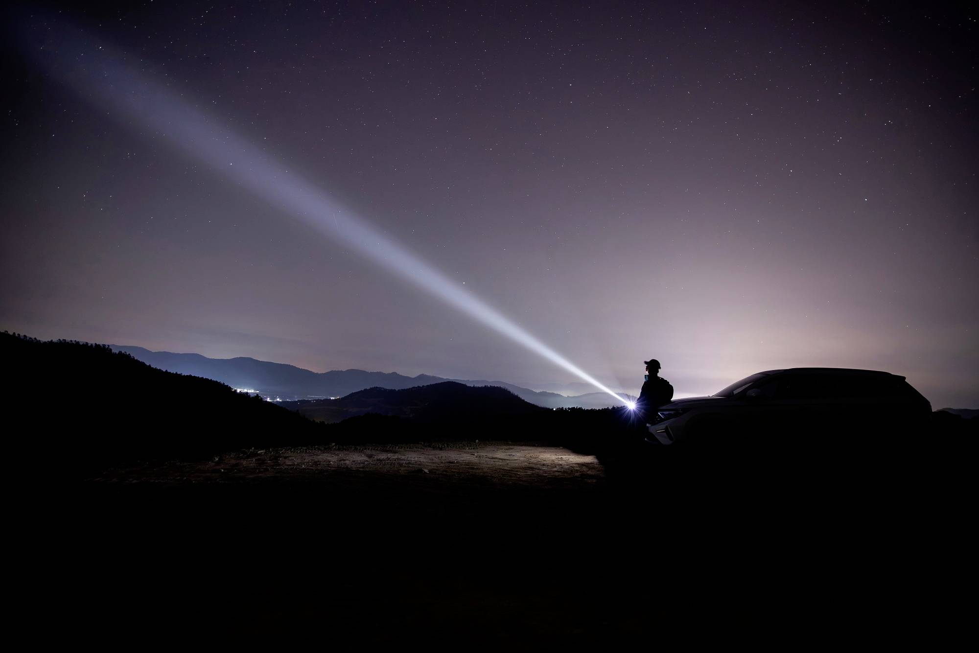 Une personne se tient la nuit à côté d'une voiture dont le phare est allumé, projetant un large faisceau lumineux dans le ciel sombre.