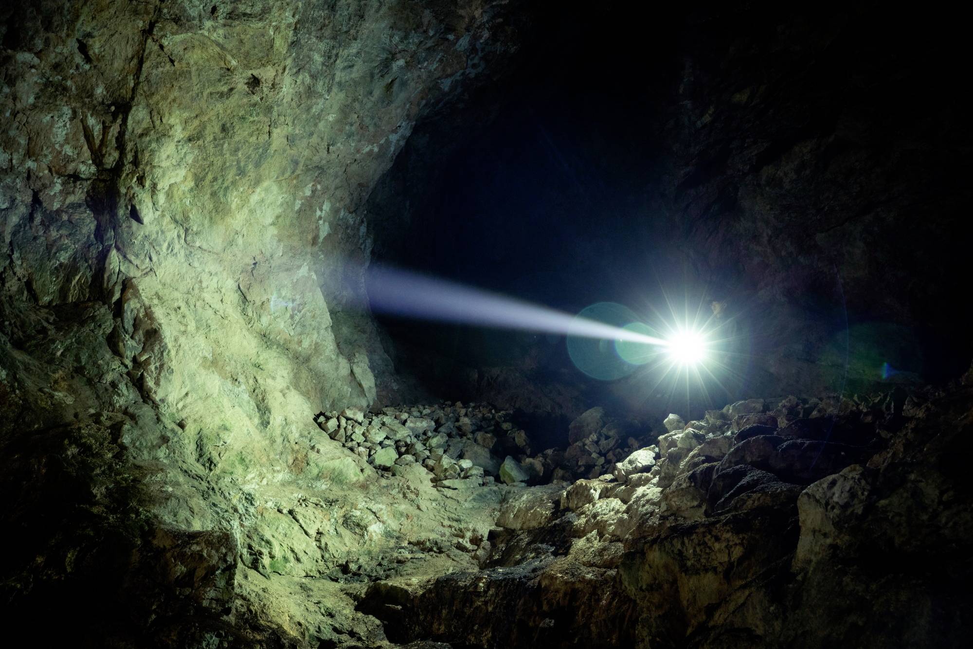 Un rayon de lumière vive pénètre dans une grotte sombre, révélant des parois rocheuses et un sol irrégulier.